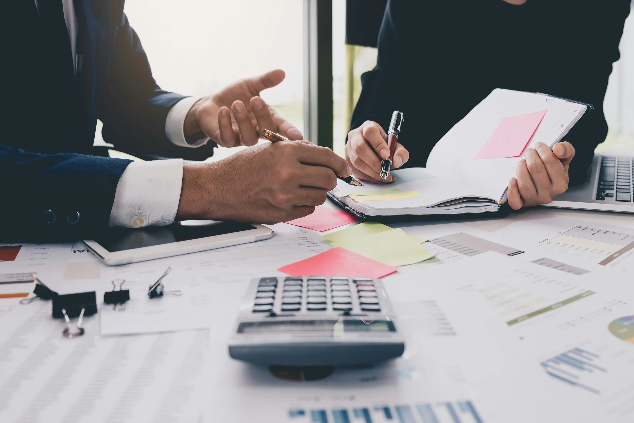 People in business attire exchanging a handshake at a cluttered desk with papers, sticky notes, a laptop, and office supplies.