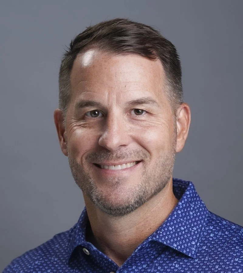 Headshot of a smiling man with short dark hair and a beard, wearing a blue collared shirt, against a gray background.