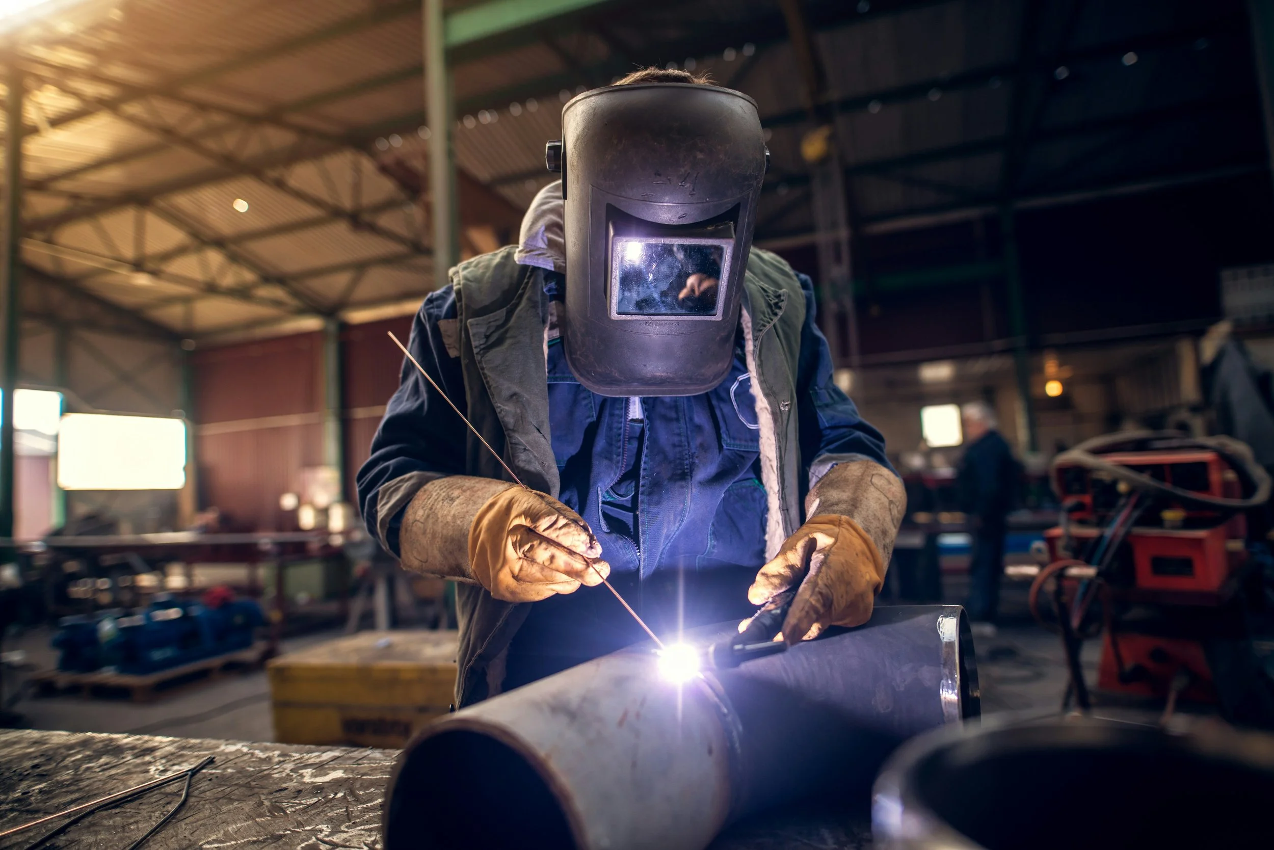 Worker welding metal pipe in an industrial workshop, wearing protective gear including a welding helmet and gloves.