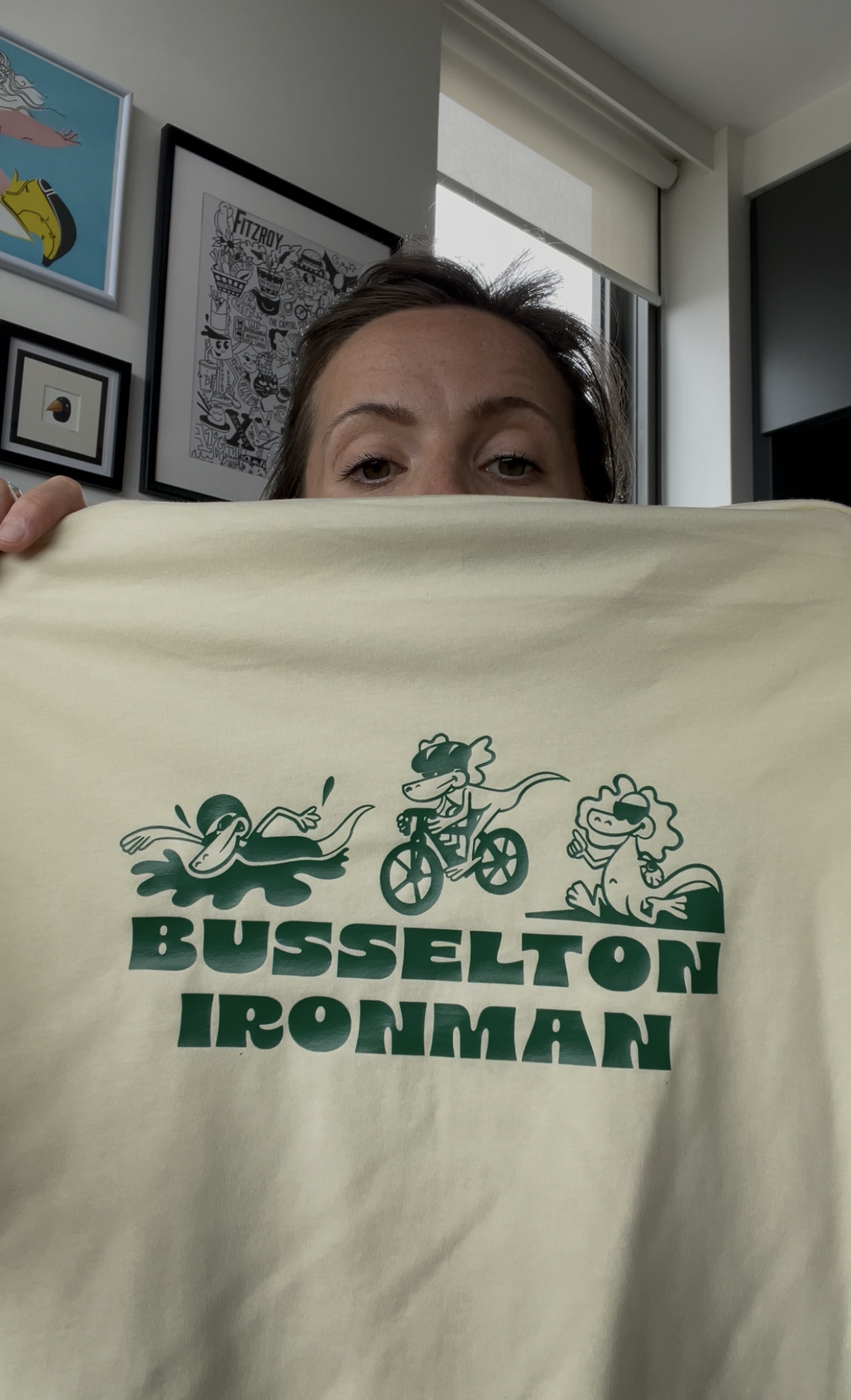 A woman holding a yellow shirt with the logo of Busselton Ironman, featuring cartoon characters of a swimmer, cyclist, and runner, and the words 'Busselton Ironman' underneath.