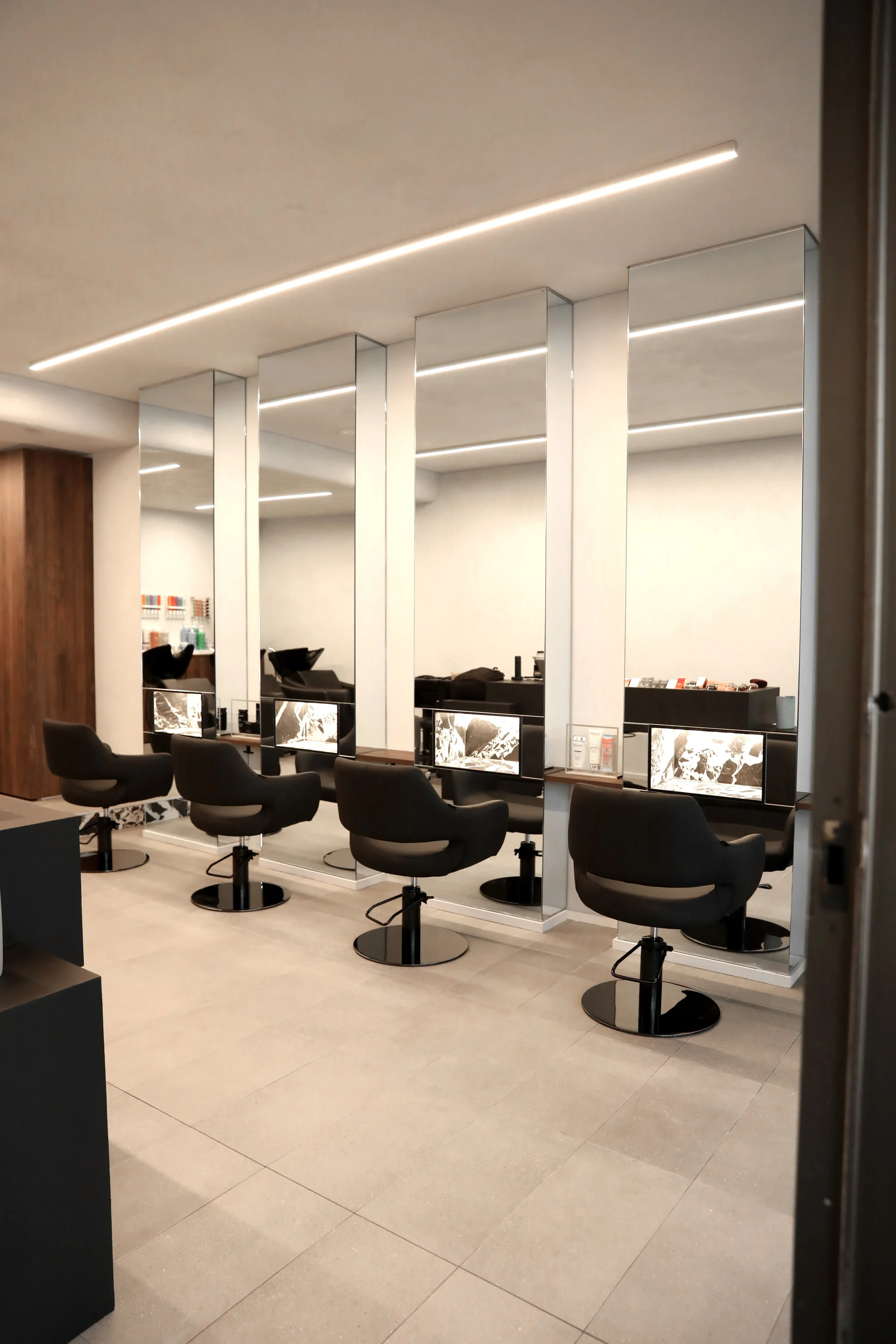 Interior view of a modern hair salon with black styling chairs, mirrors, and hair products on shelves. The ceiling has sleek, curved LED lighting, and the floors are light-colored tiles.