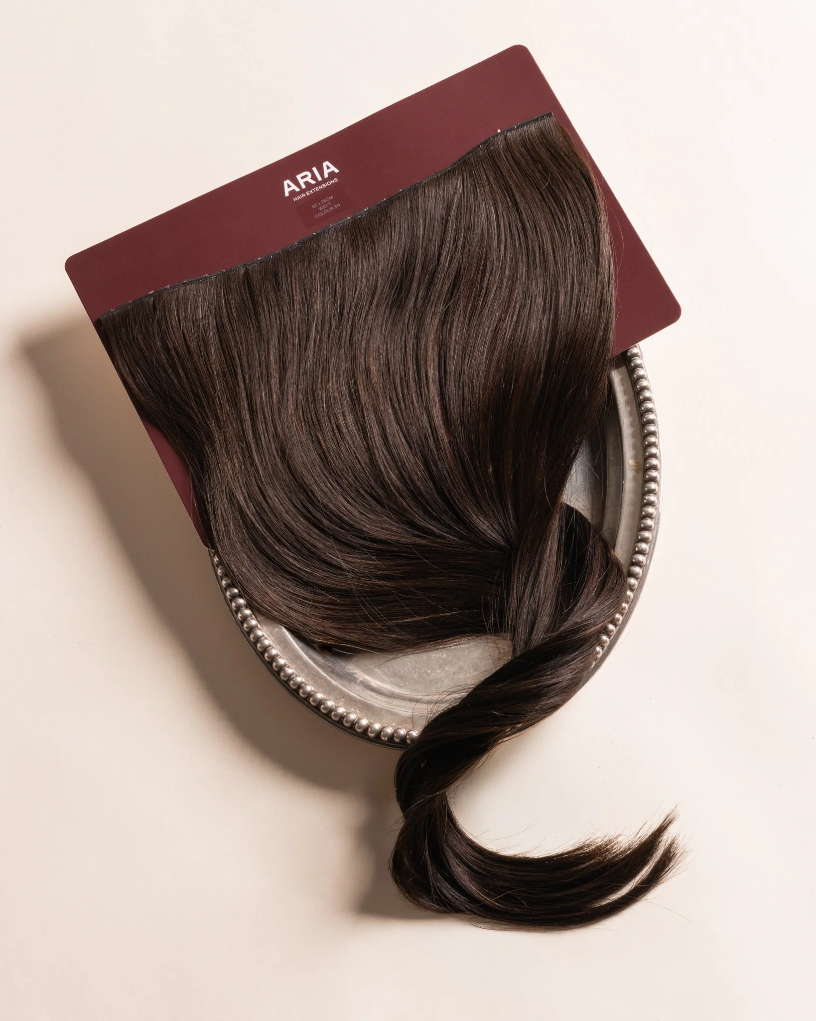 Butterfly Weft hair extensions on ARIA packaging box with white text, placed on a silver tray with beaded edging. The box reads 'ARIA HAIR EXTENSIONS'.