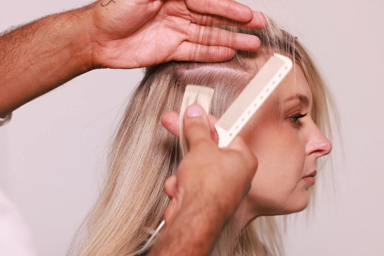 A person installing tape-in hair extensions into a blonde woman's hair