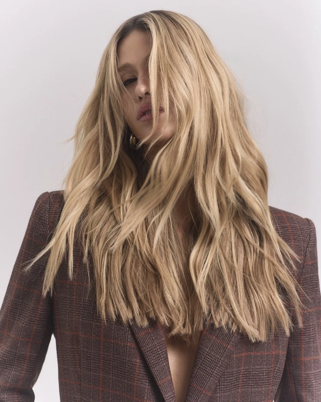 A woman with long, wavy blonde hair, partially covering her face, wearing a plaid blazer against a plain white background.