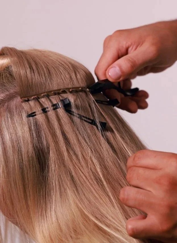 A person is installing Butterfly Weft hair extensions into a person’s blonde hair, with sections of hair separated and clipped.