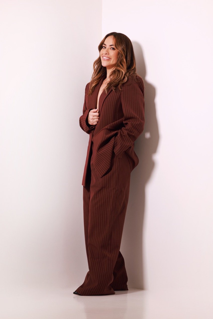 A woman with long, wavy brown hair wearing a burgundy pinstripe suit, standing against a plain white wall, smiling and looking at the camera.