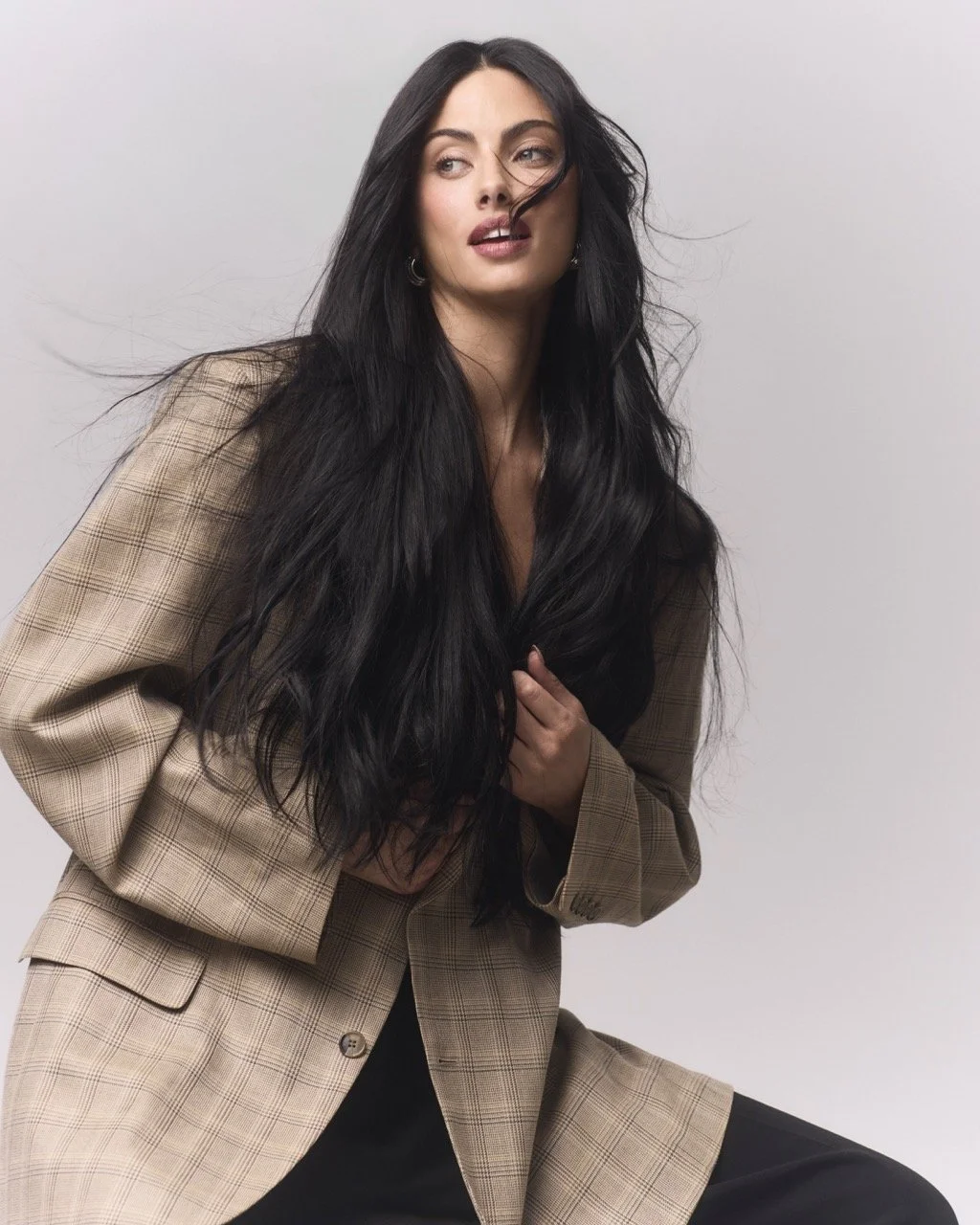 A woman with long black hair wearing a beige plaid blazer over a black top, posing against a plain white background.