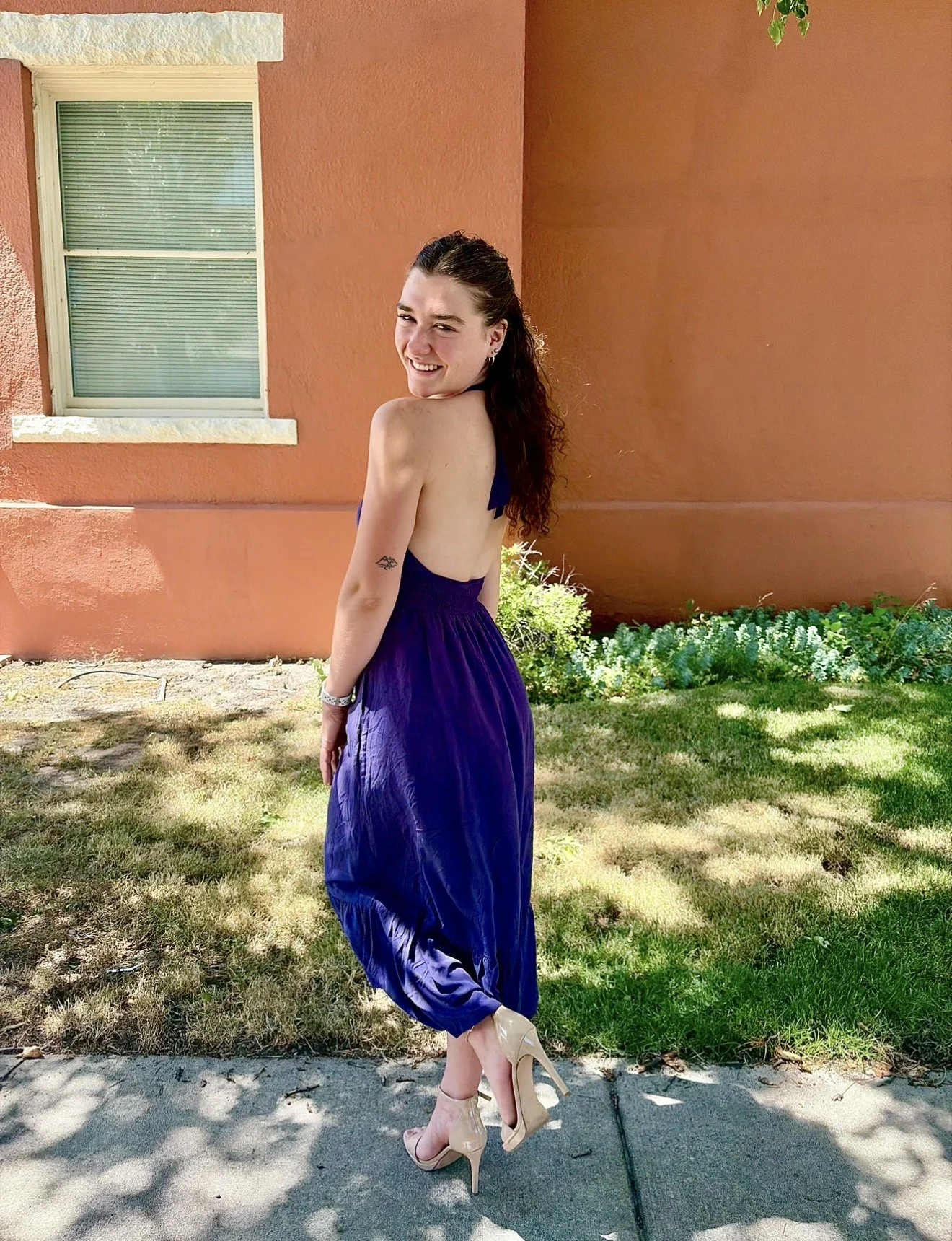 A young woman with long dark hair smiling and posing outdoors in front of a pink wall with a window, wearing a sleeveless purple dress and nude high heels, standing on grass and concrete.