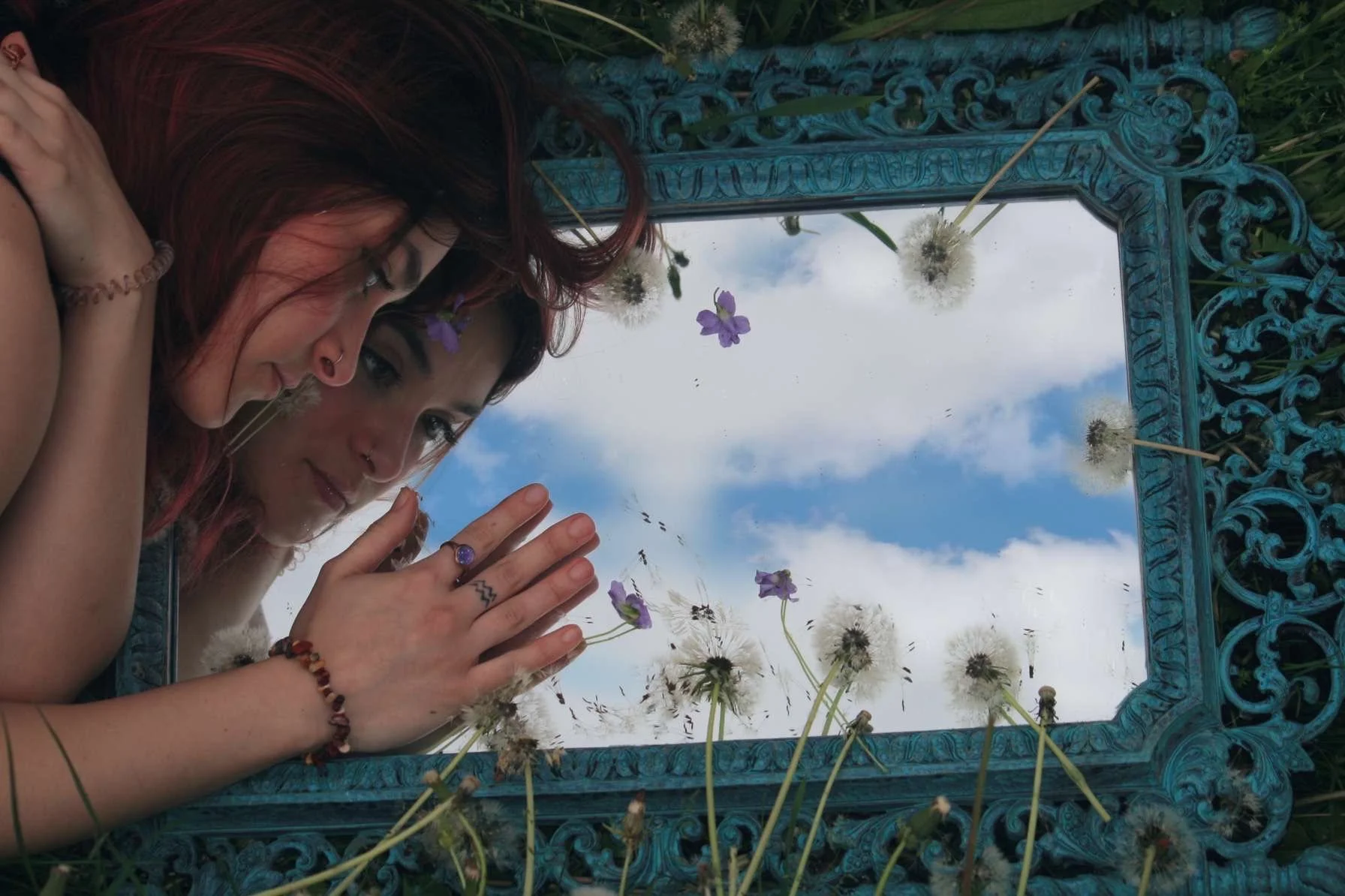 Two women are lying on the ground next to a reflective mirror with an ornate blue frame, surrounded by dandelions and purple flowers, looking into the mirror with sky and clouds reflected in it.