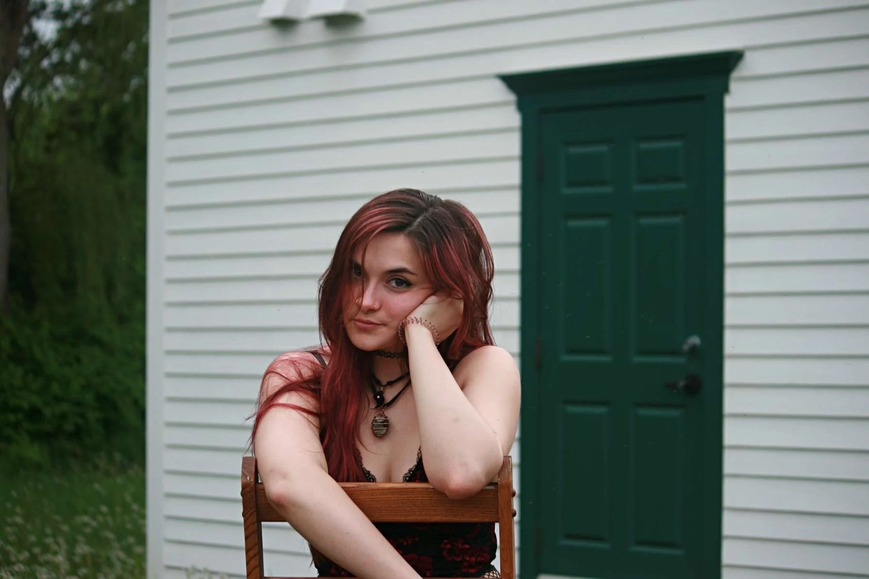 A young woman with long red hair and piercings sits outdoors, resting her head on her hand, in front of a white wooden building with a green door.