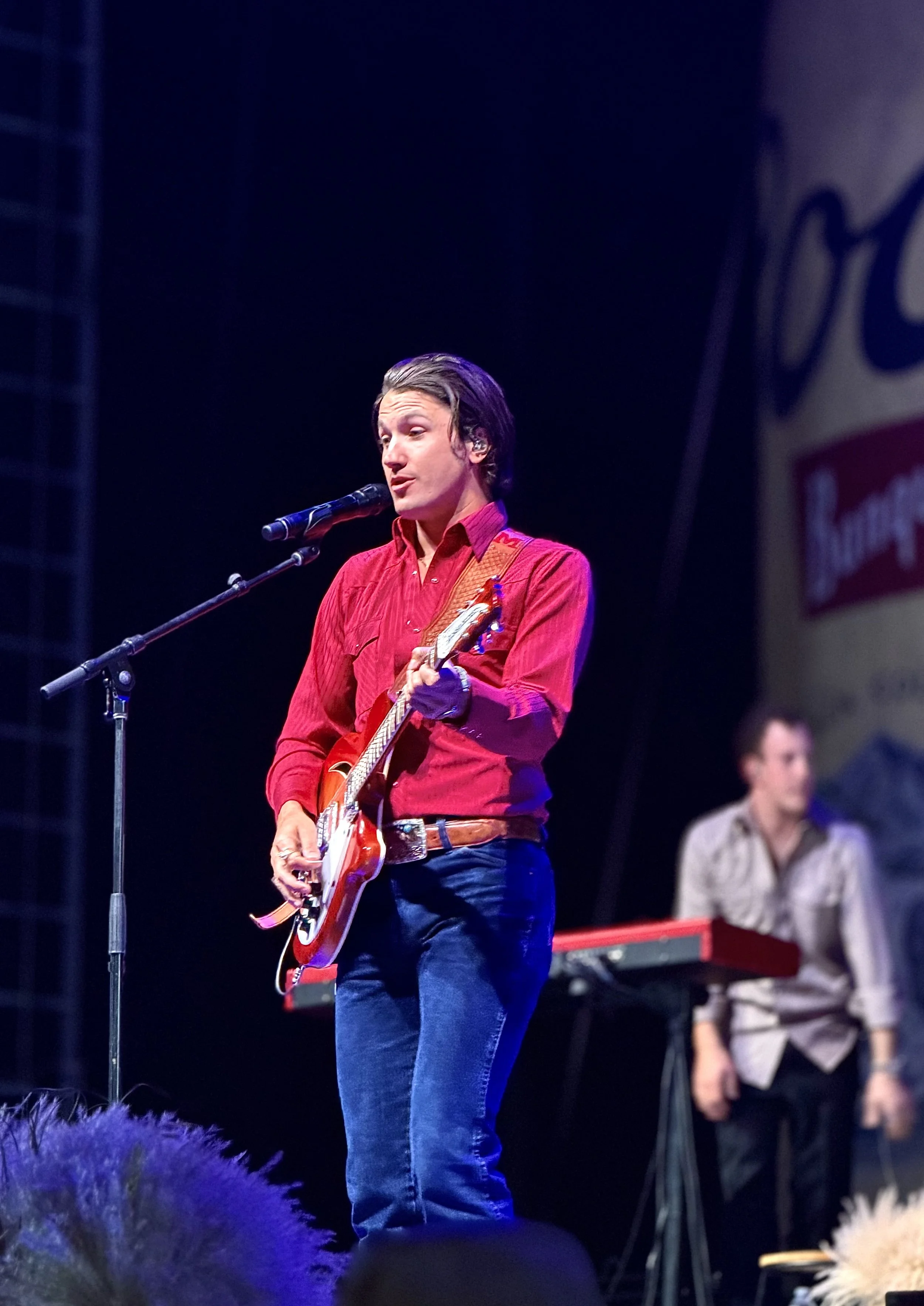 A male musician wearing a red shirt and jeans, playing an electric guitar and singing into a microphone on stage. Two other musicians are in the background, one playing a keyboard and the other playing a bass guitar.