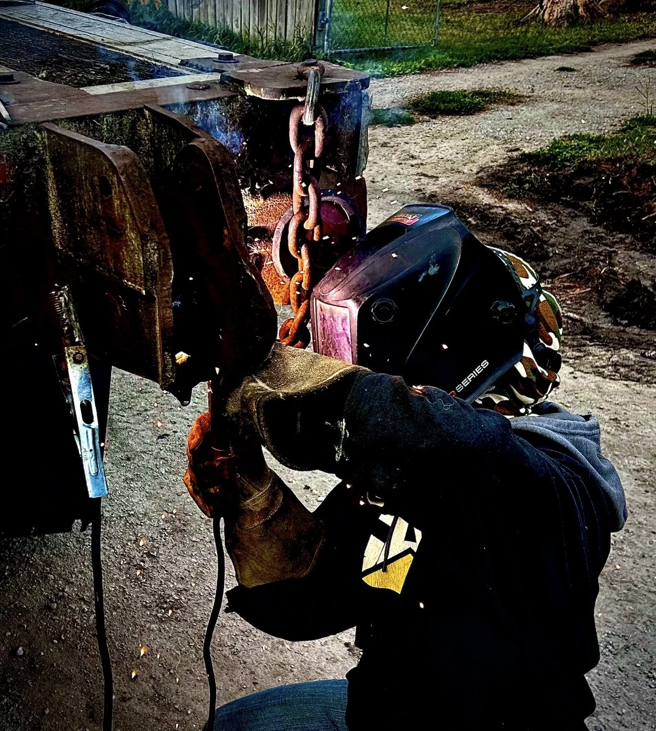 A person wearing a welding helmet and gloves is welding a large metal structure outdoors, with sparks flying from the welding process.