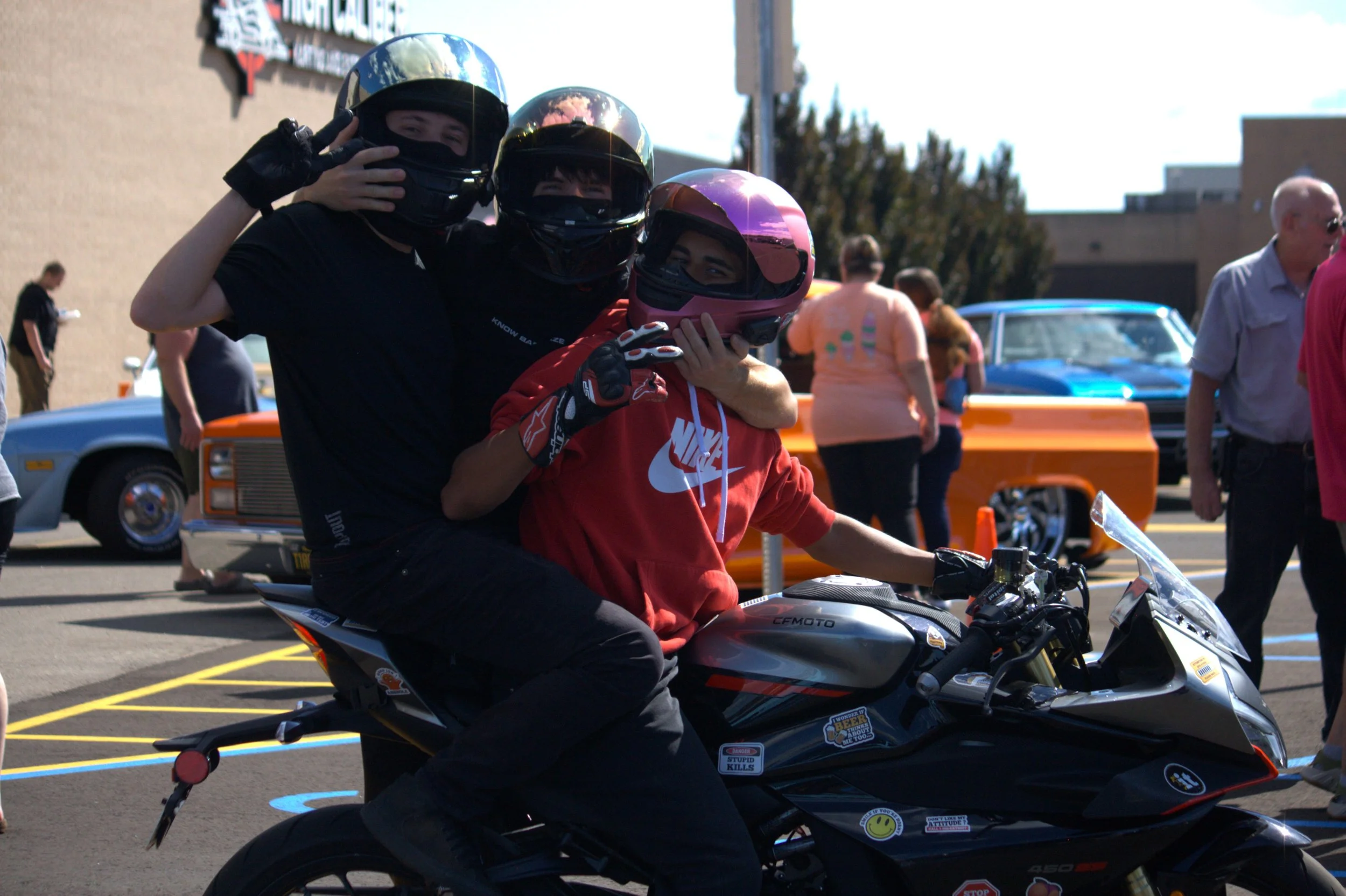 Three people on motorcycles wearing helmets, surrounded by people and vintage cars in an outdoor setting.
