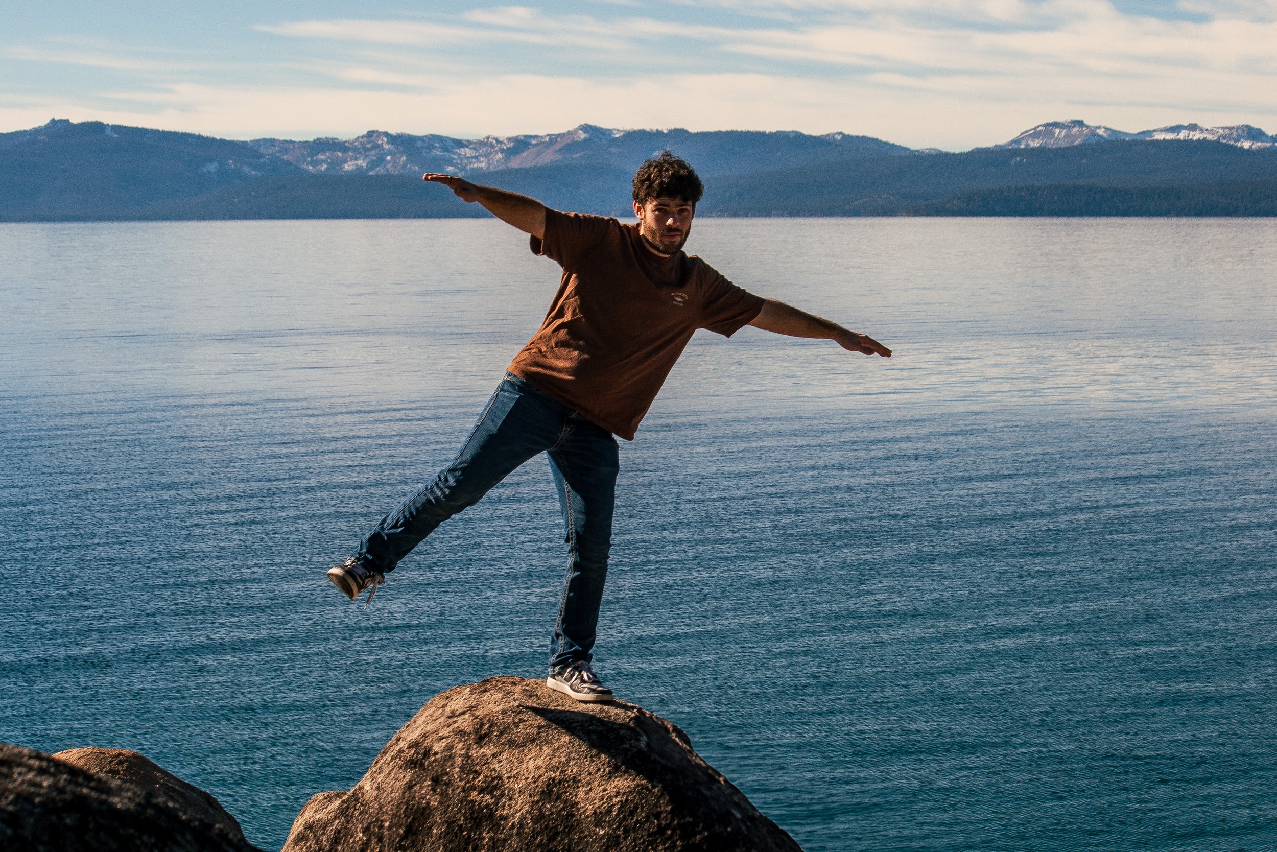 A man balancing on a large rock in the water, with mountains and a cloudy sky in the background.