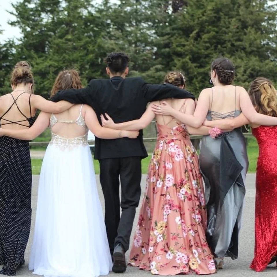 Group of six young women and one young man walking outdoors, seen from the back, with arms around each other. The women are wearing colorful evening dresses and the man is in a black suit. The background features green trees and grass, suggesting a park or garden setting.