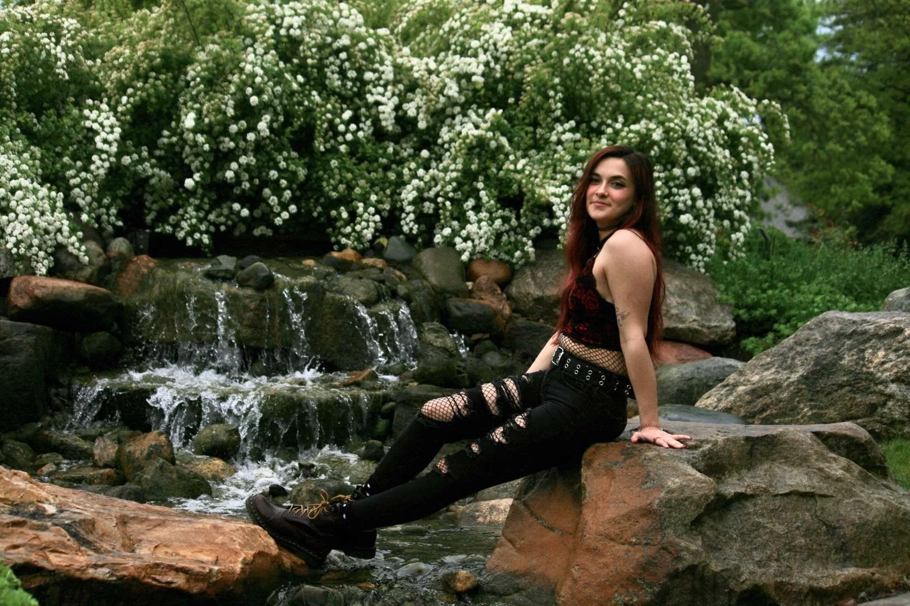 A young woman with long dark red hair sitting on rocks by a small waterfall in a lush green setting with white flowers in the background.