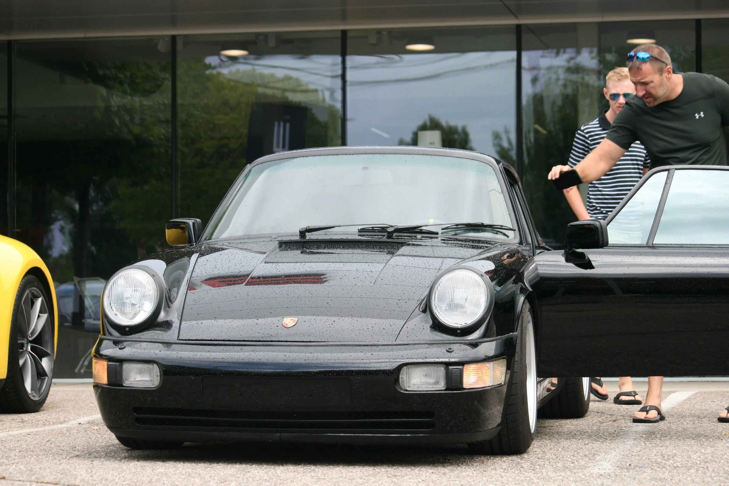 A black Porsche sports car parked outside a glass building, with two men examining the vehicle. One man is wearing sunglasses and a black shirt, while the other is in a striped shirt.