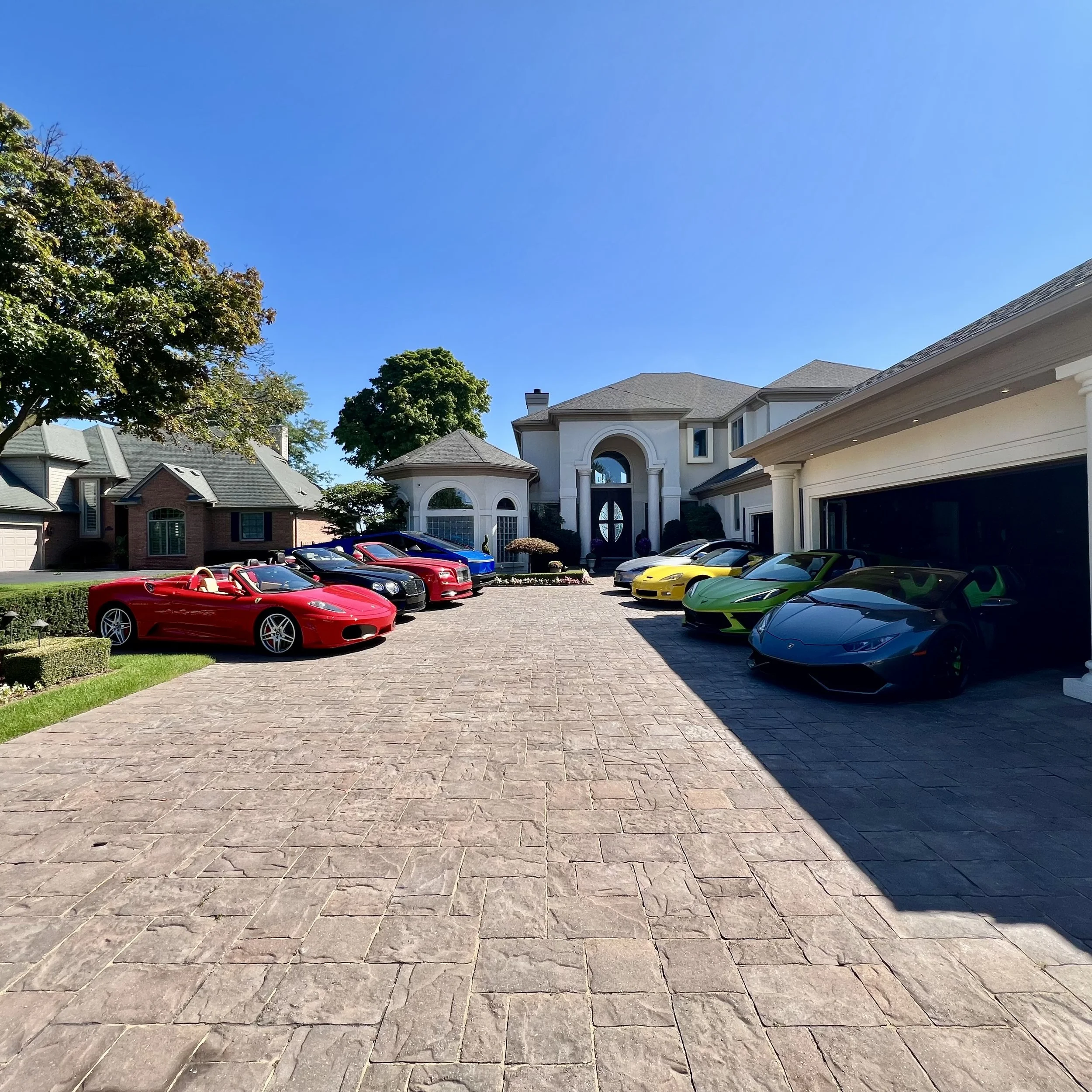 View of a residential neighborhood with large houses, a driveway with multiple sports cars, including red, yellow, green, and black vehicles, and a clear blue sky.