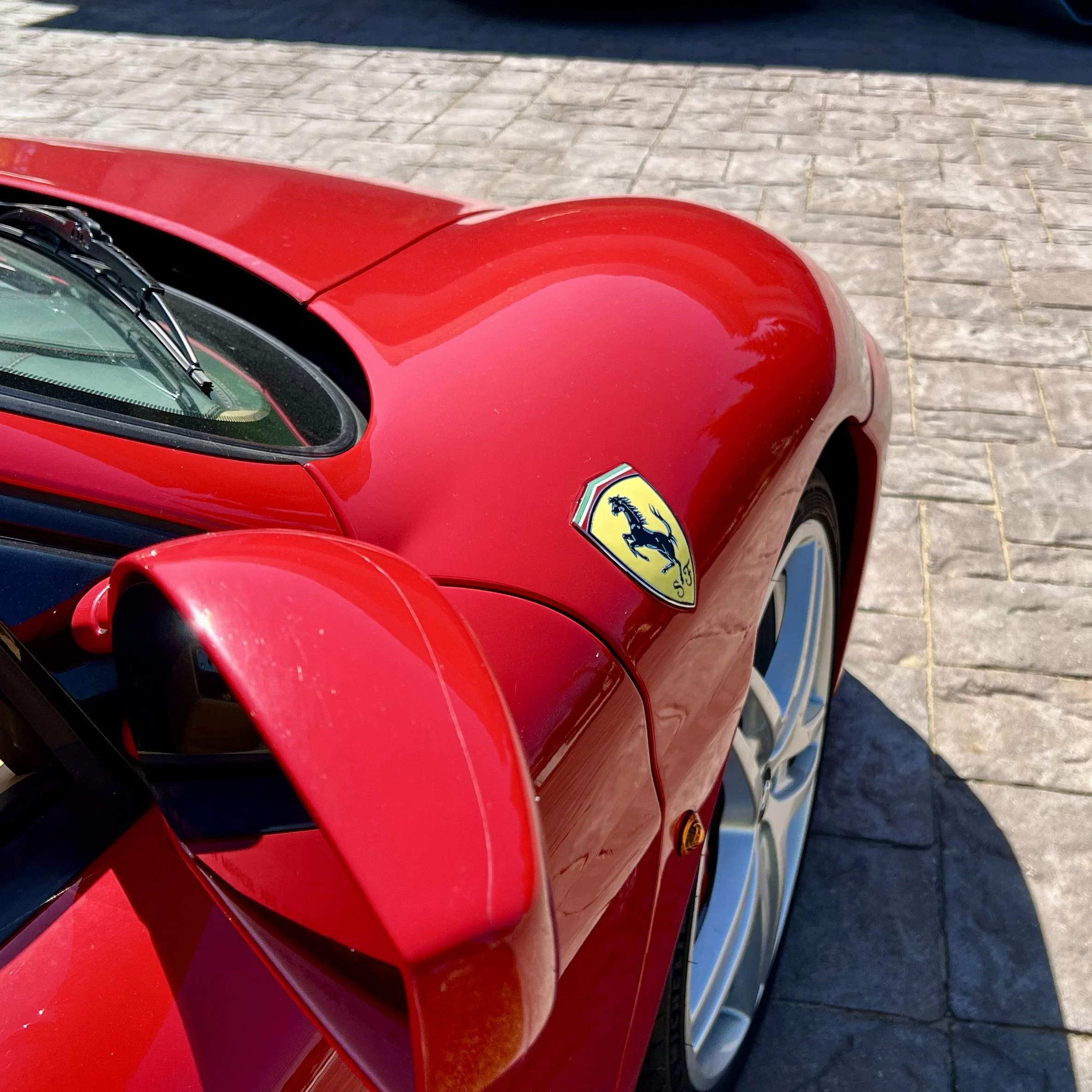 Close-up of the front part of a red Ferrari sports car parked on a stone-paved street, showing the Ferrari badge on the hood.