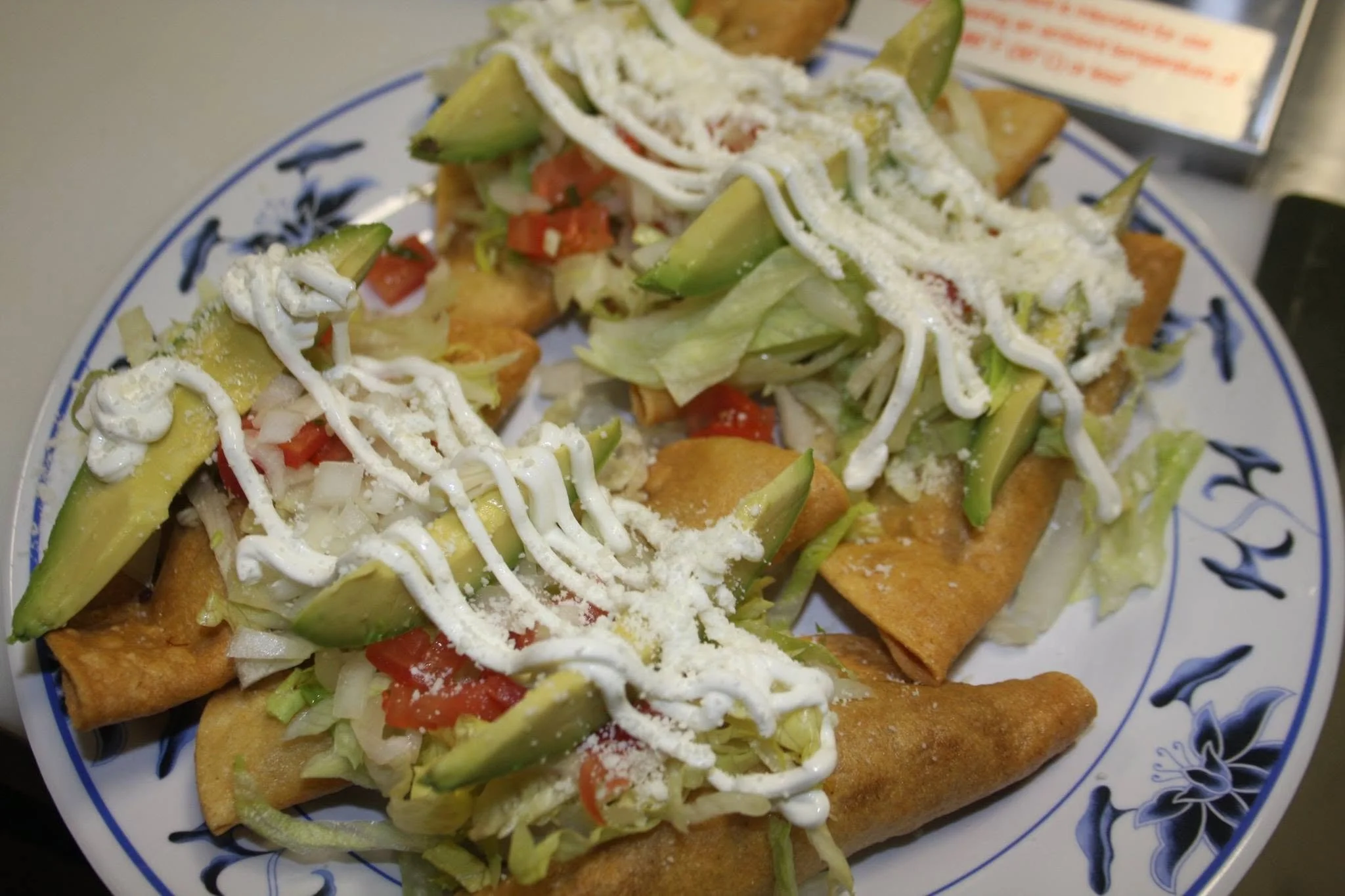 Close-up of a plate of fish tacos topped with diced tomatoes, shredded lettuce, sliced avocados, shredded cheese, and drizzled with sour cream, served on a decorative blue and white plate.
