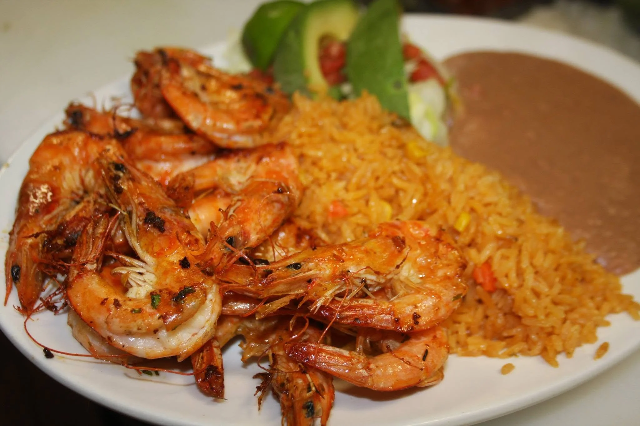 Close-up of a plate with grilled shrimp, yellow rice, sliced avocado, and brown sauce on the side.