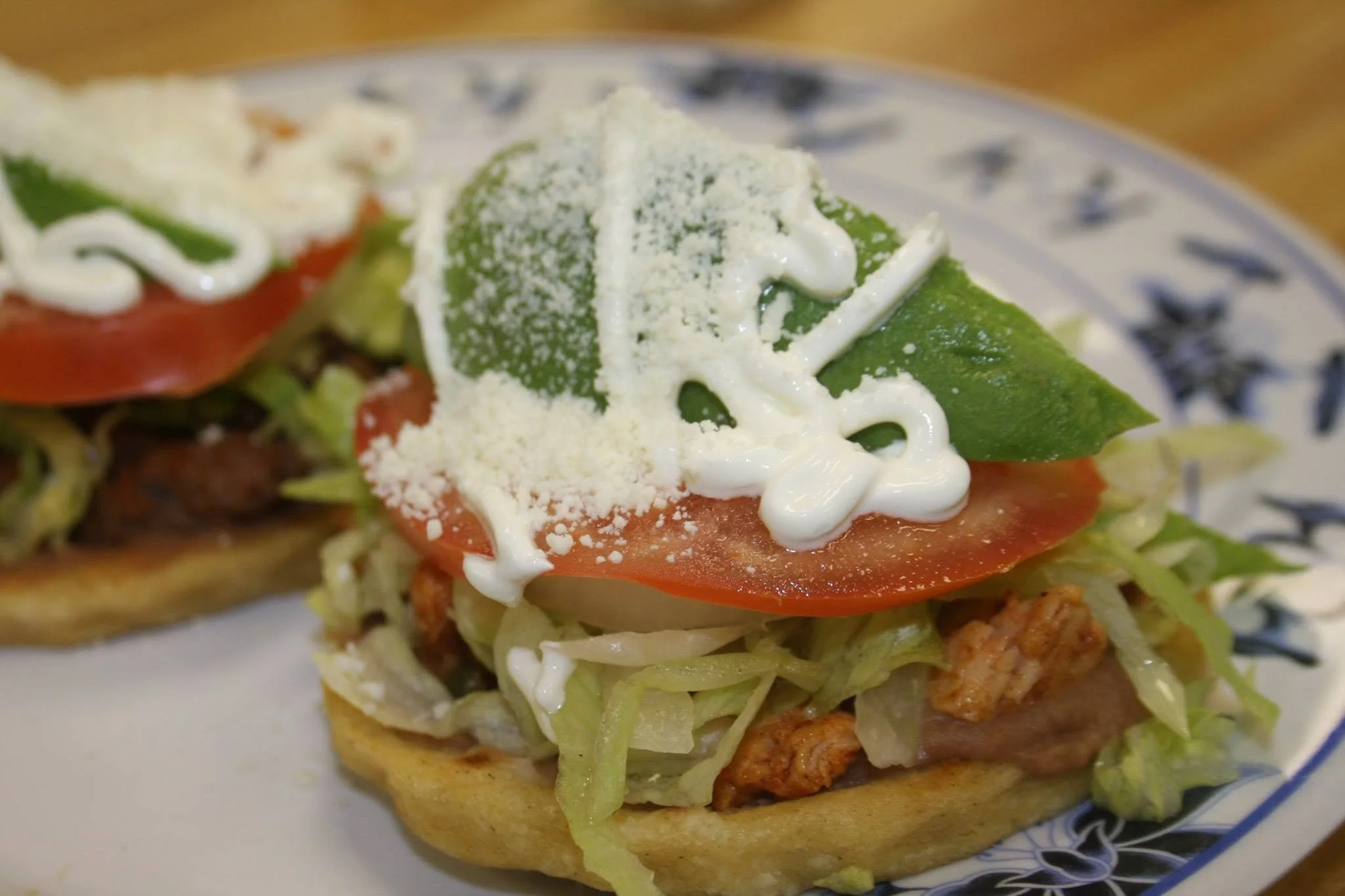 Close-up of a taco with lettuce, tomato, creamy white sauce, grated cheese, and a green jalapeño on top, served on a decorative plate.