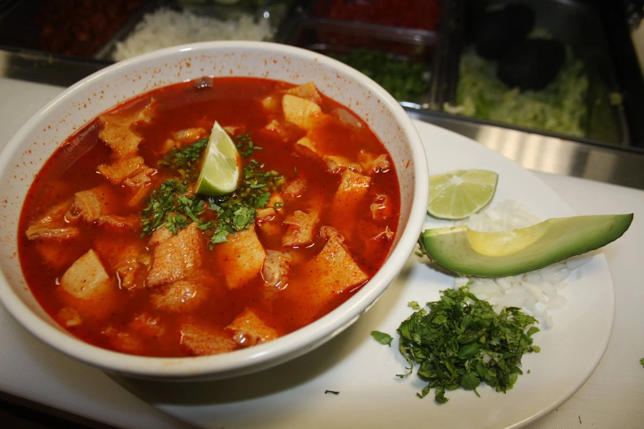A bowl of red Mexican soup garnished with a lime wedge and chopped cilantro, on a white plate with additional lime and cilantro for garnish.