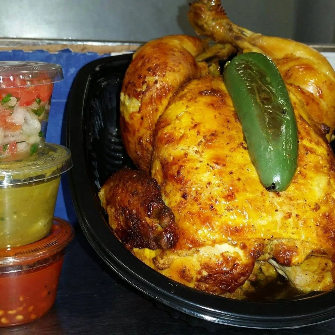 Roast whole chicken with a jalapeño placed on top, served in a black tray, with small containers of salsa and relish on the side.