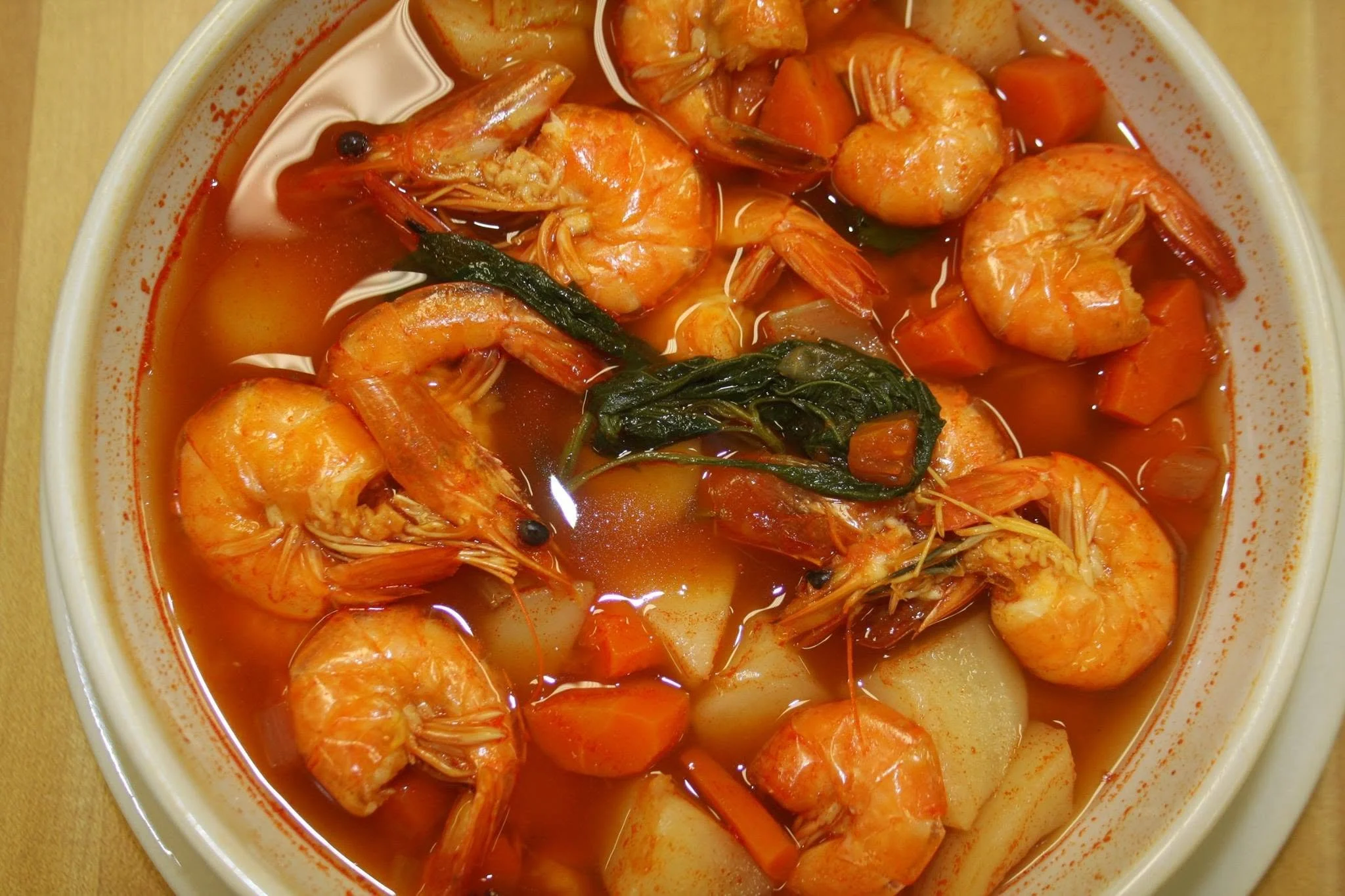 A bowl of shrimp and vegetable soup with a red broth, shrimp, carrots, potatoes, and greens.