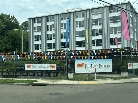 A multi-story building with colorful flags and banners in front, including signs that say "Midtown Nursery". The area appears to be decorated for a celebration or event.