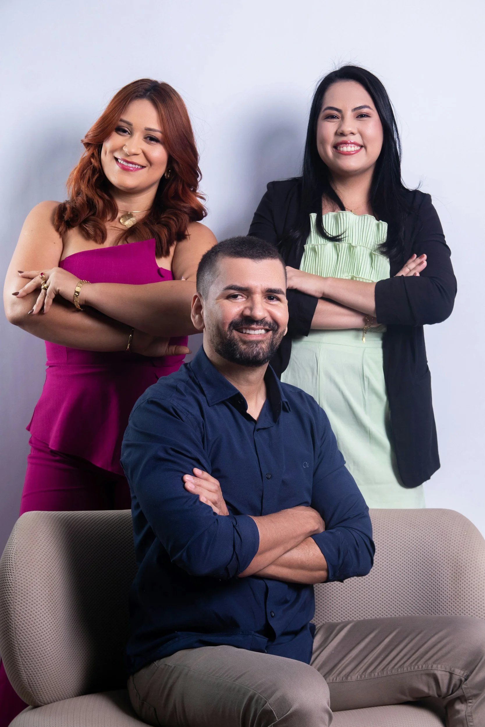 Three smiling people, two women standing behind a seated man, all with crossed arms, against a light background.