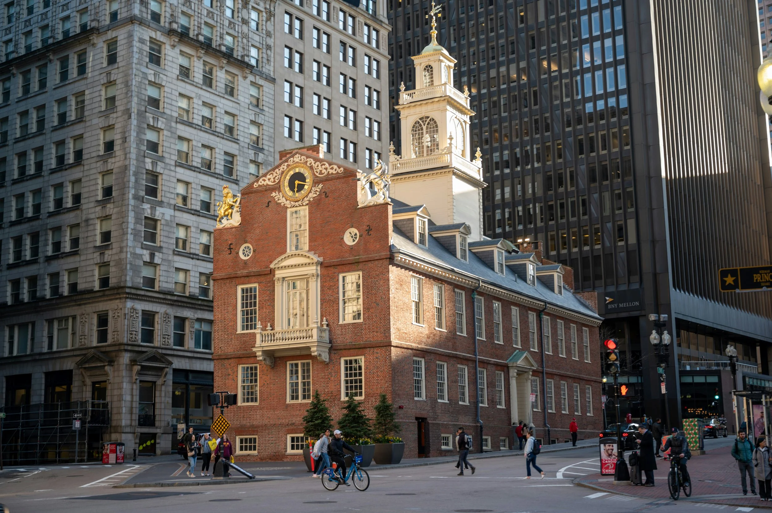 City street scene featuring a historic brick building with a clock and decorative statues, surrounded by modern high-rise buildings, pedestrians, cyclists, and street signs.