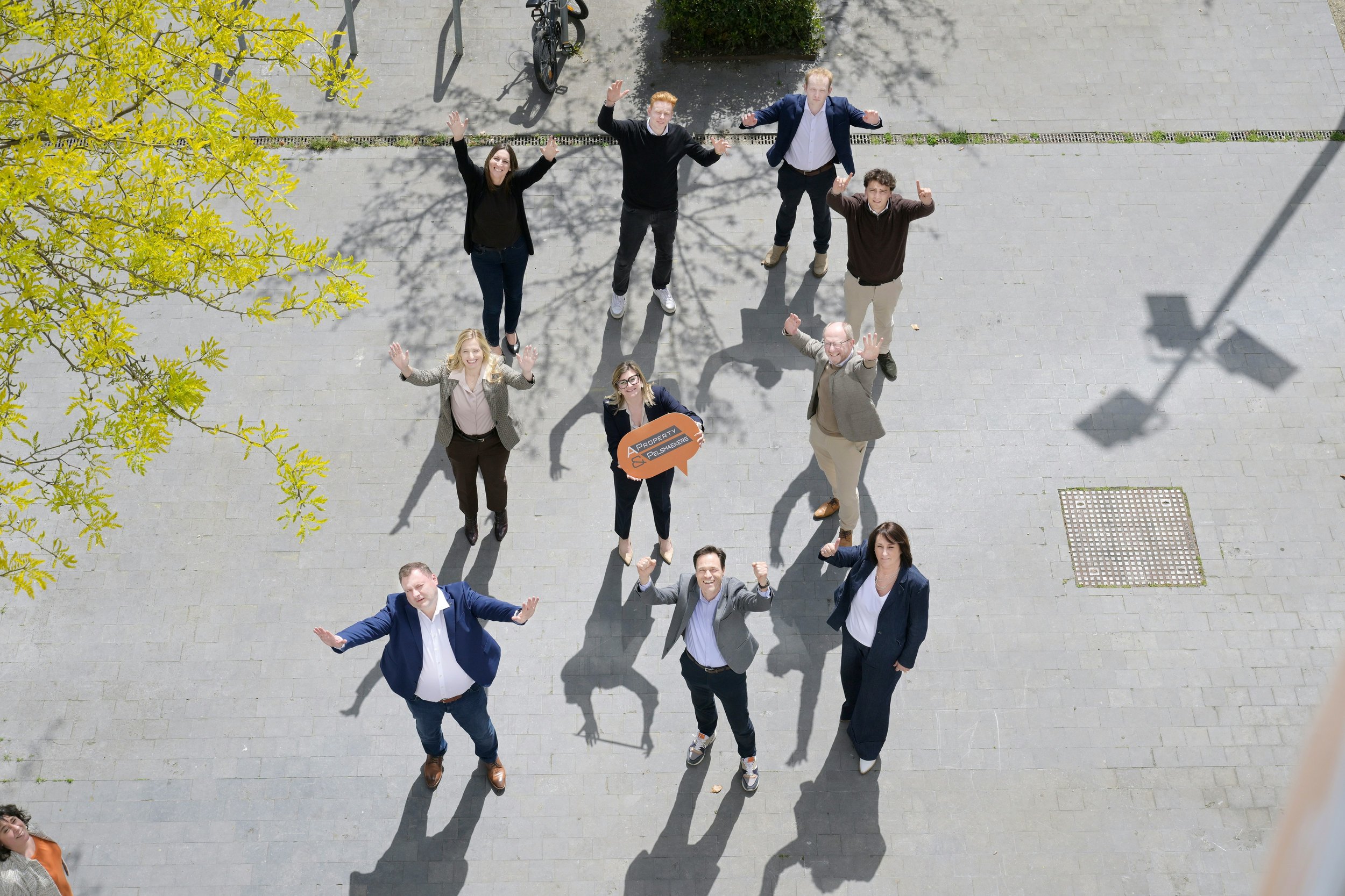 A group of people standing on a paved outdoor area, waving at the camera with their hands raised. They are smiling and appear to be posing for the photo. There are trees, a bicycle, and shadows on the ground.