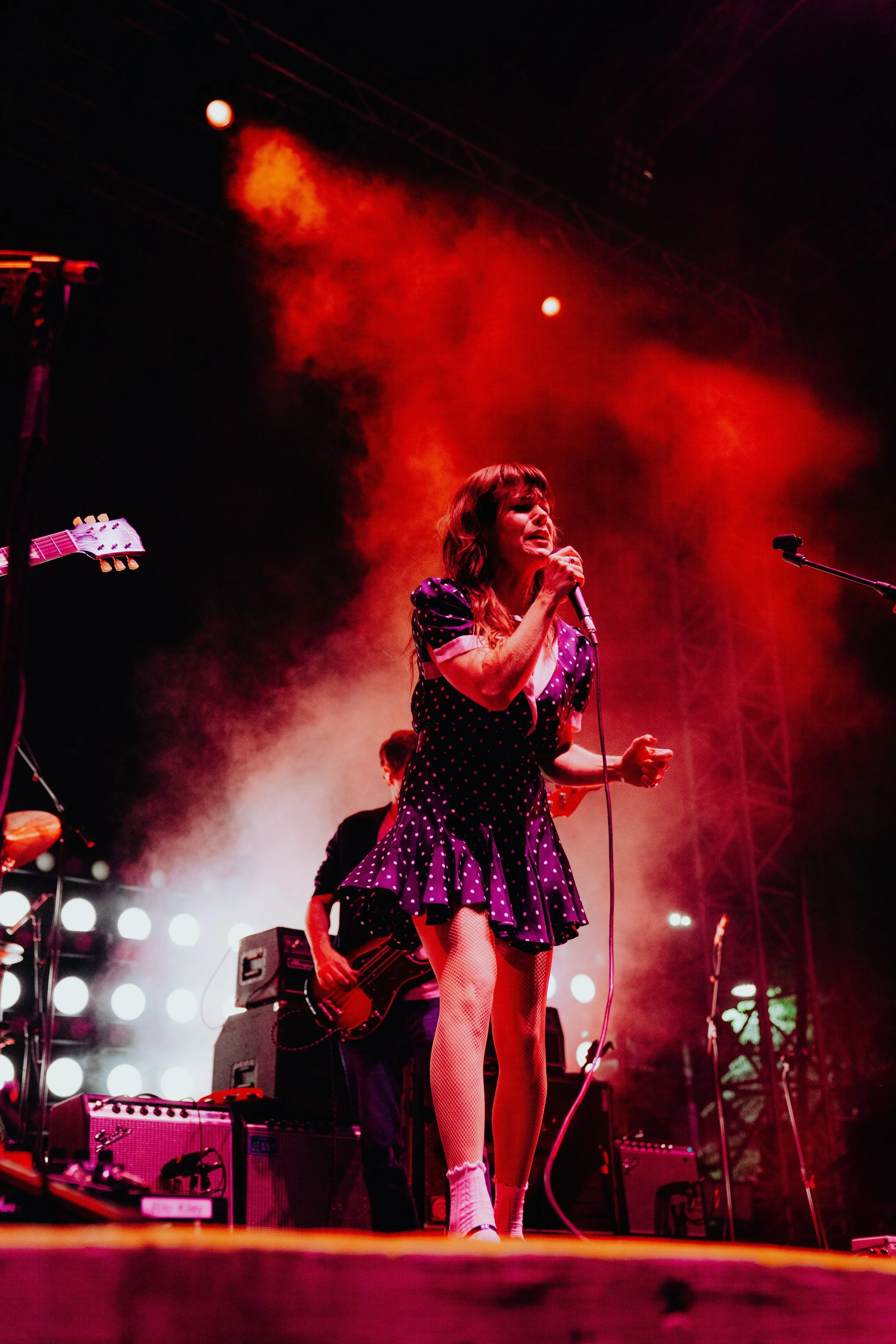 A female singer on stage wearing a purple polka dot dress with white knee-high socks singing into a microphone under red stage lighting. There are musical instruments and equipment visible in the background.