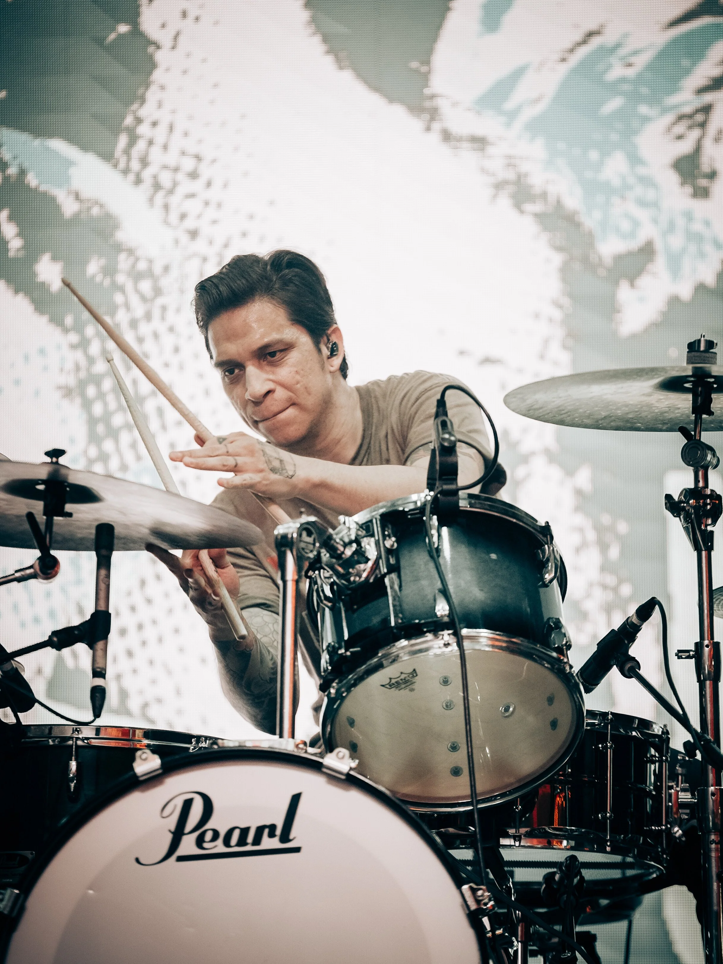 A man appears to be playing a drum set, mainly a snare drum and a cymbal, with a serious expression. He is wearing a beige shirt and has tape on his left forearm. The background features a wall with an abstract or pixelated pattern.
