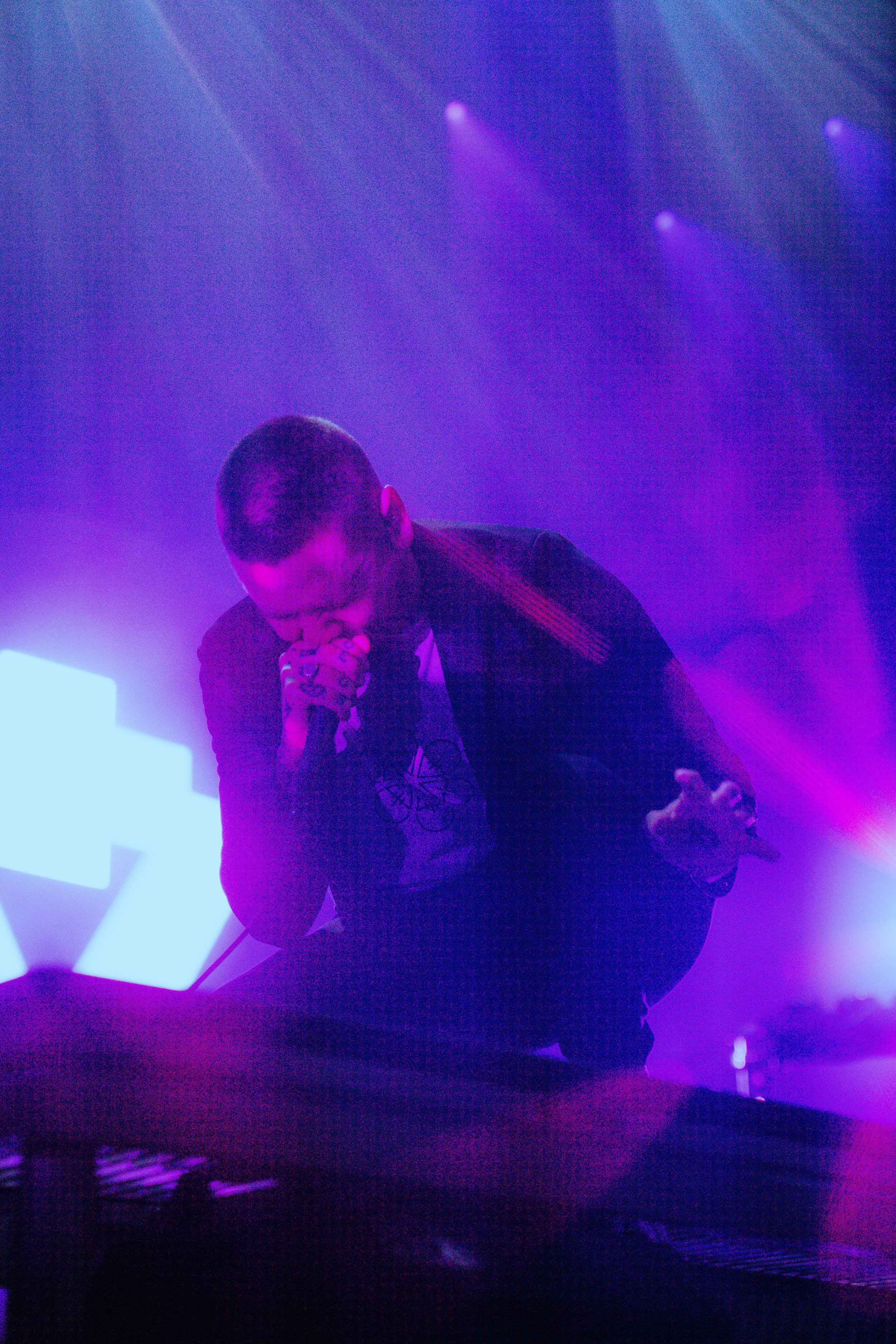 A male performer with short dark hair singing into a microphone on a stage illuminated with purple and blue lights.