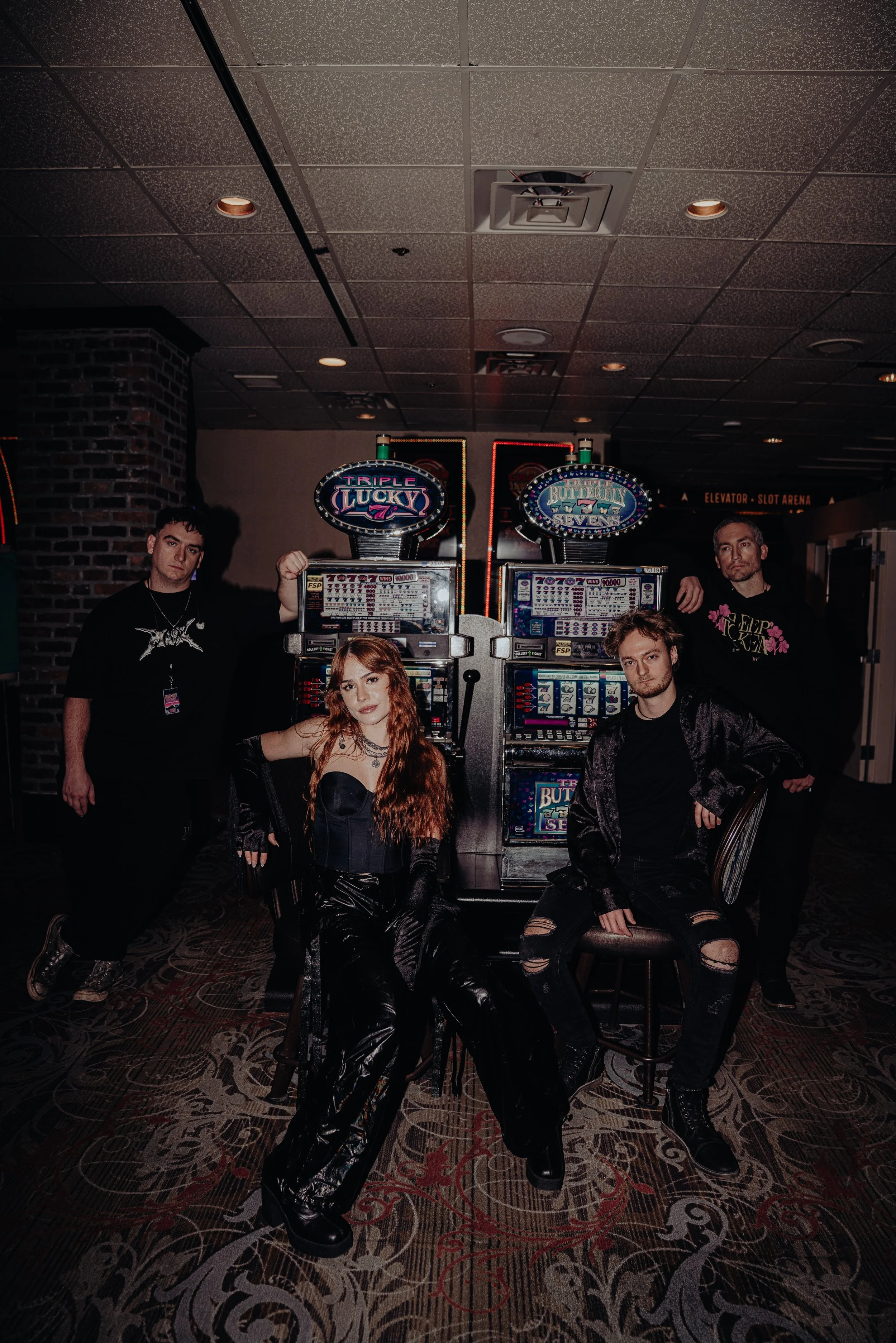 A group of five young adults posing in front of slot machines at a casino. Four males and one female. The female has long, curly red hair and is wearing a black strapless top with matching pants. The males are dressed in dark, casual clothing, with t