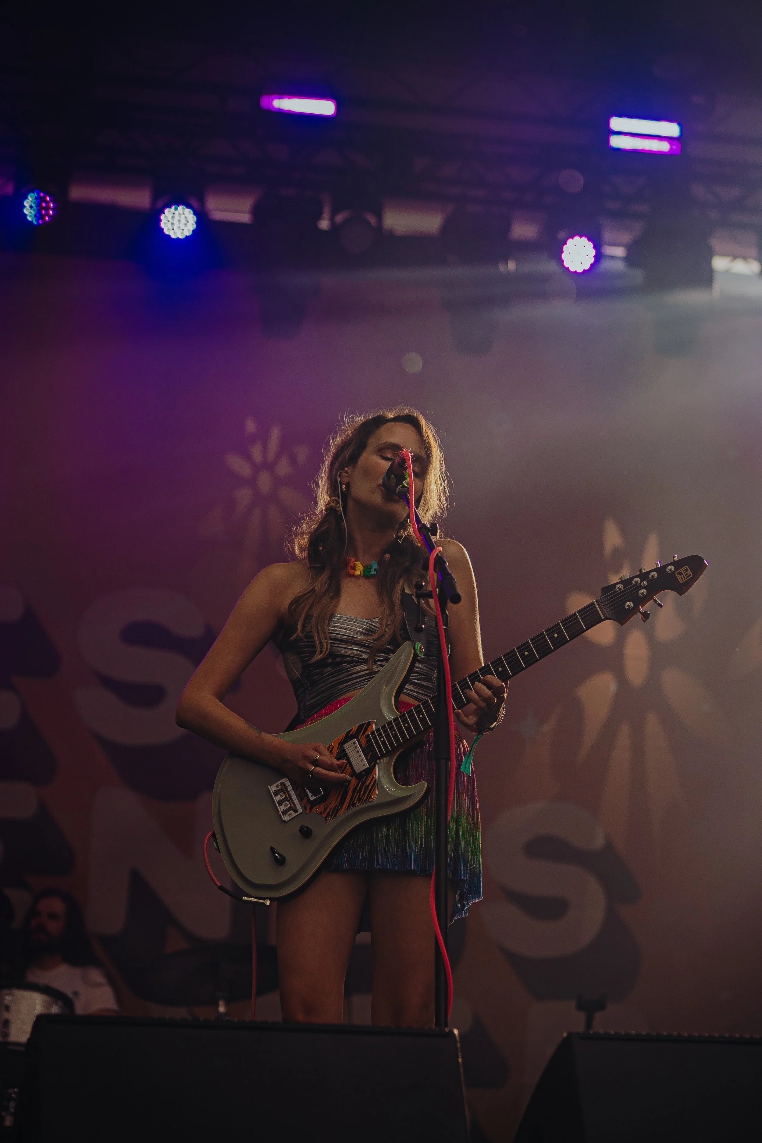 A woman with long, wavy hair playing an electric guitar on stage during a concert. She is wearing a colorful dress and appears to be singing into a microphone. Stage lights are illuminating her and the background features a patterned banner.
