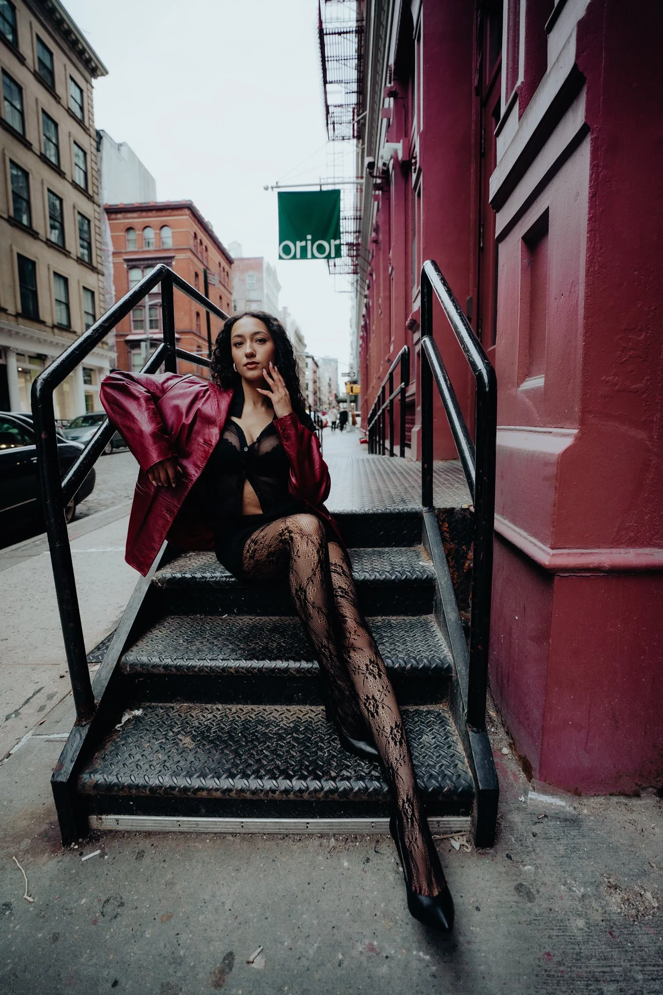 A woman with curly black hair sitting on metal stairs outside a red building on a city street. She is wearing a red leather jacket, a black lace outfit, fishnet stockings, and black high heels. The background includes buildings, parked cars, and an o