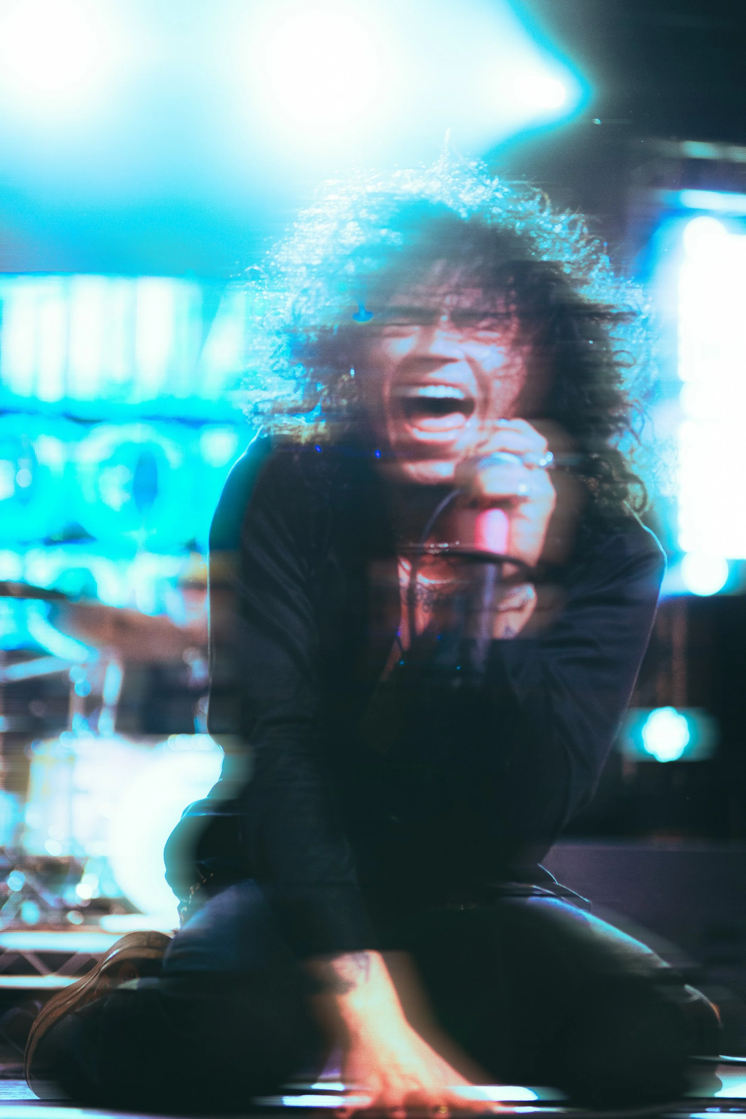 A woman with curly hair passionately singing into a microphone on a stage with bright blue and white lighting.