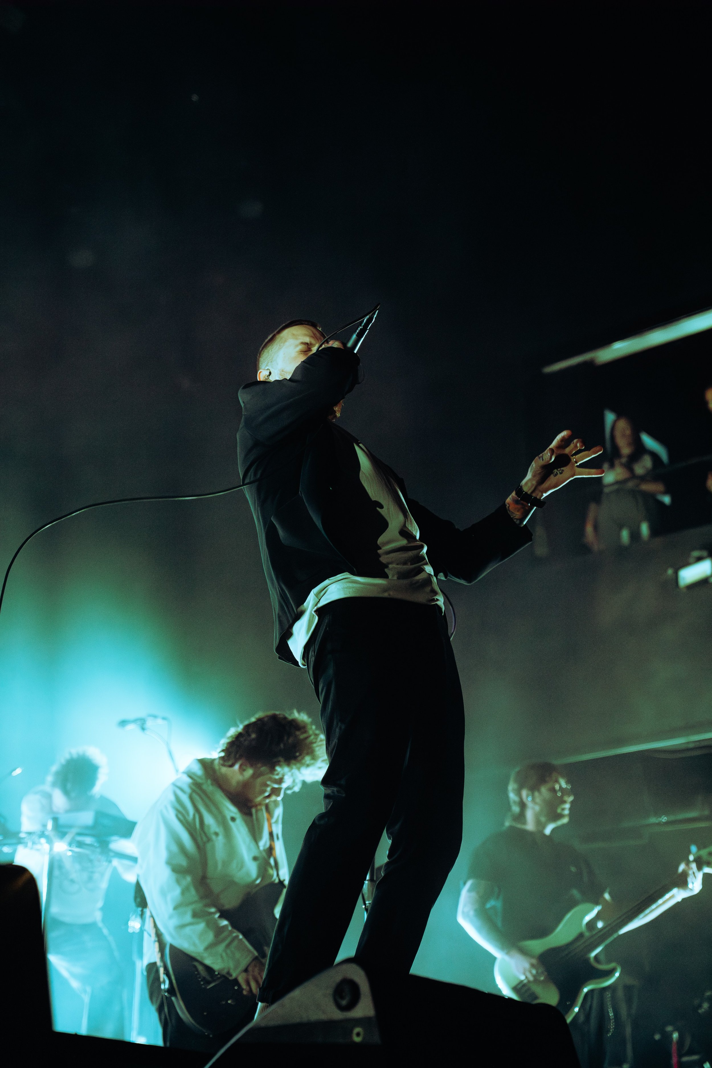 A band performs on stage in a dimly lit setting, featuring a vocalist holding a microphone, several guitarists, and a keyboard player, with colorful stage lighting.