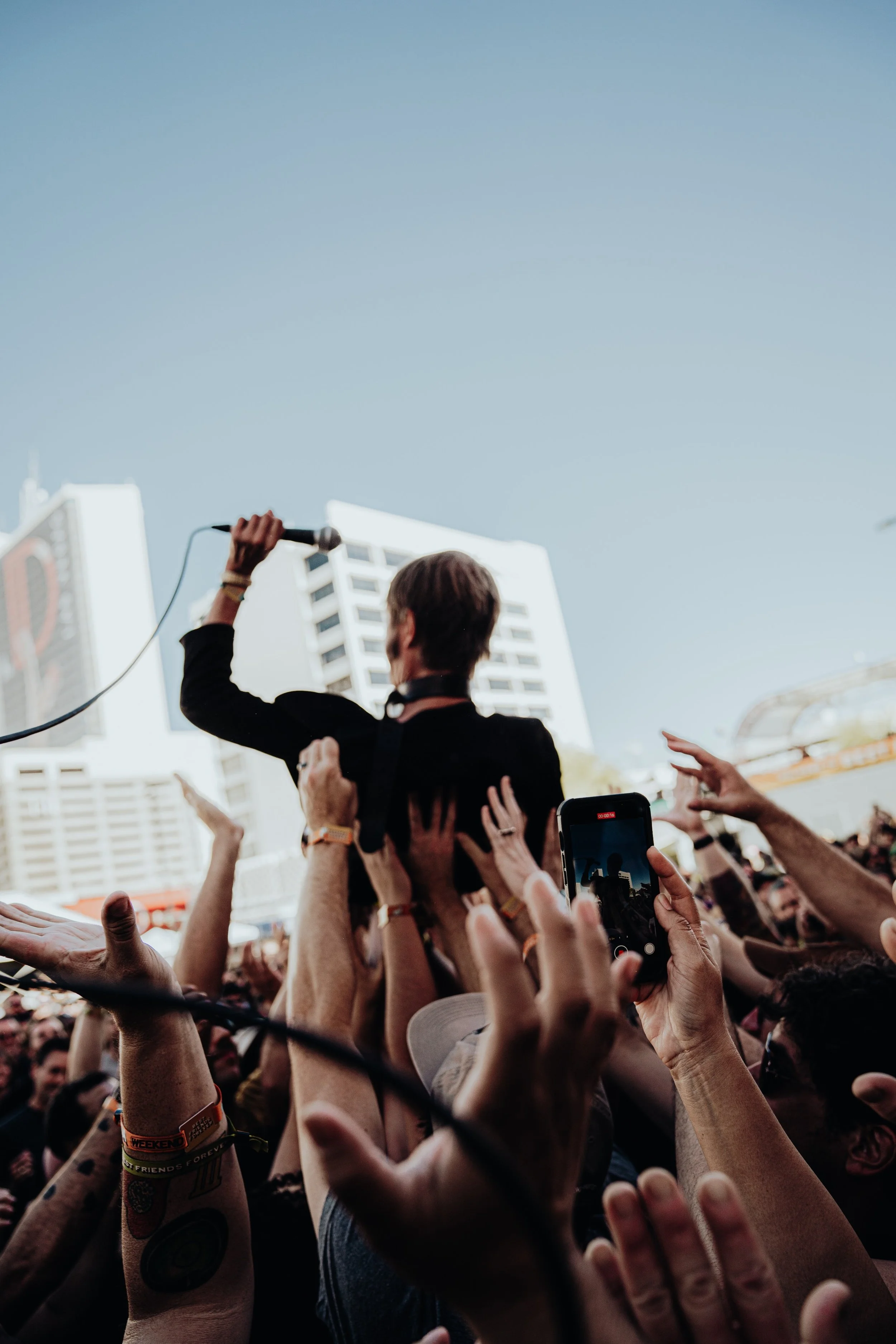 A crowd of people with their hands raised towards a performer at an outdoor event in a city. The performer is holding a microphone and facing away from the camera, with tall buildings in the background.