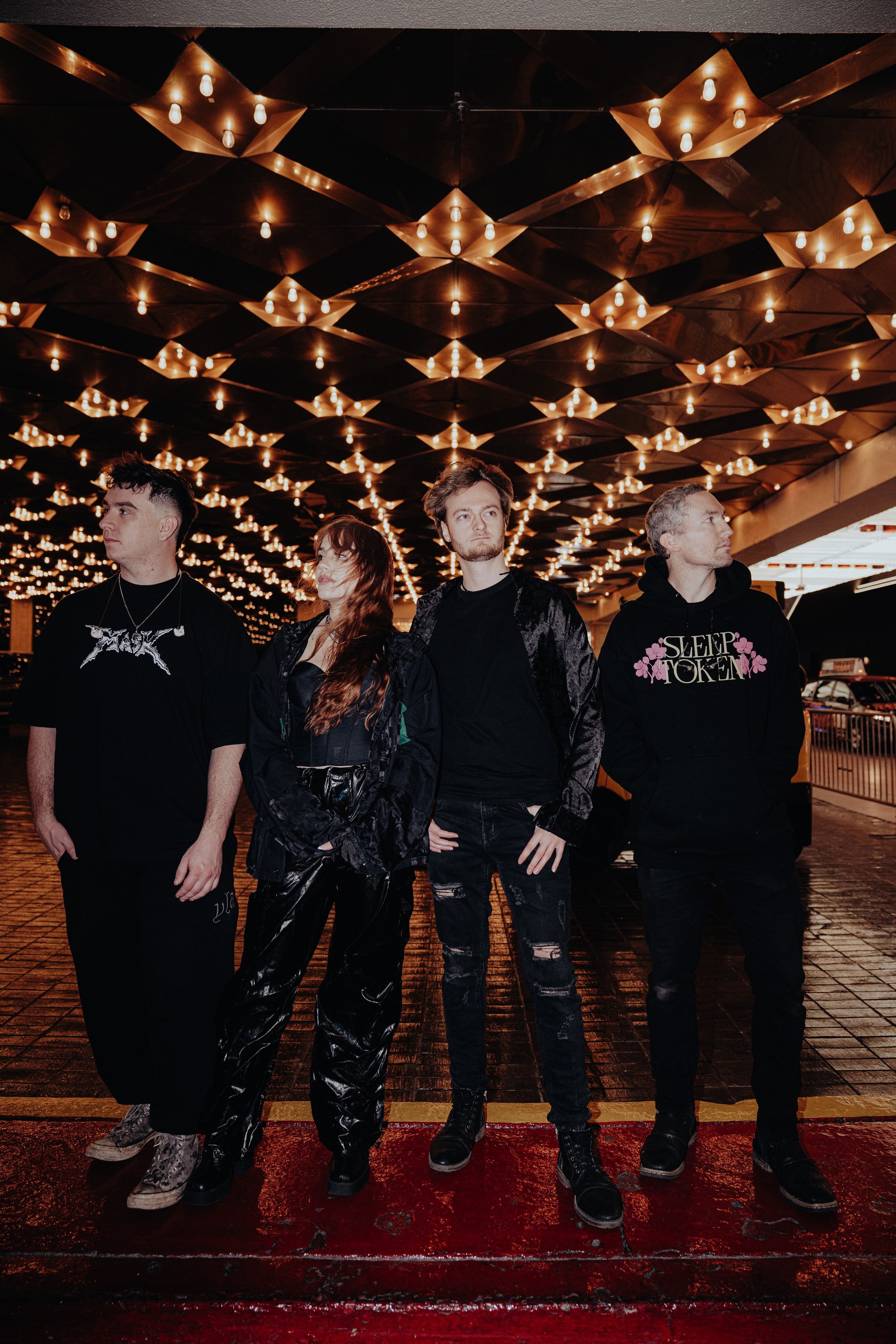 Four young adults standing under a ceiling with many small lights, at night. The group appears to be in an urban setting, possibly outside a venue or restaurant. They are dressed in dark, casual clothing with a punk or goth style.