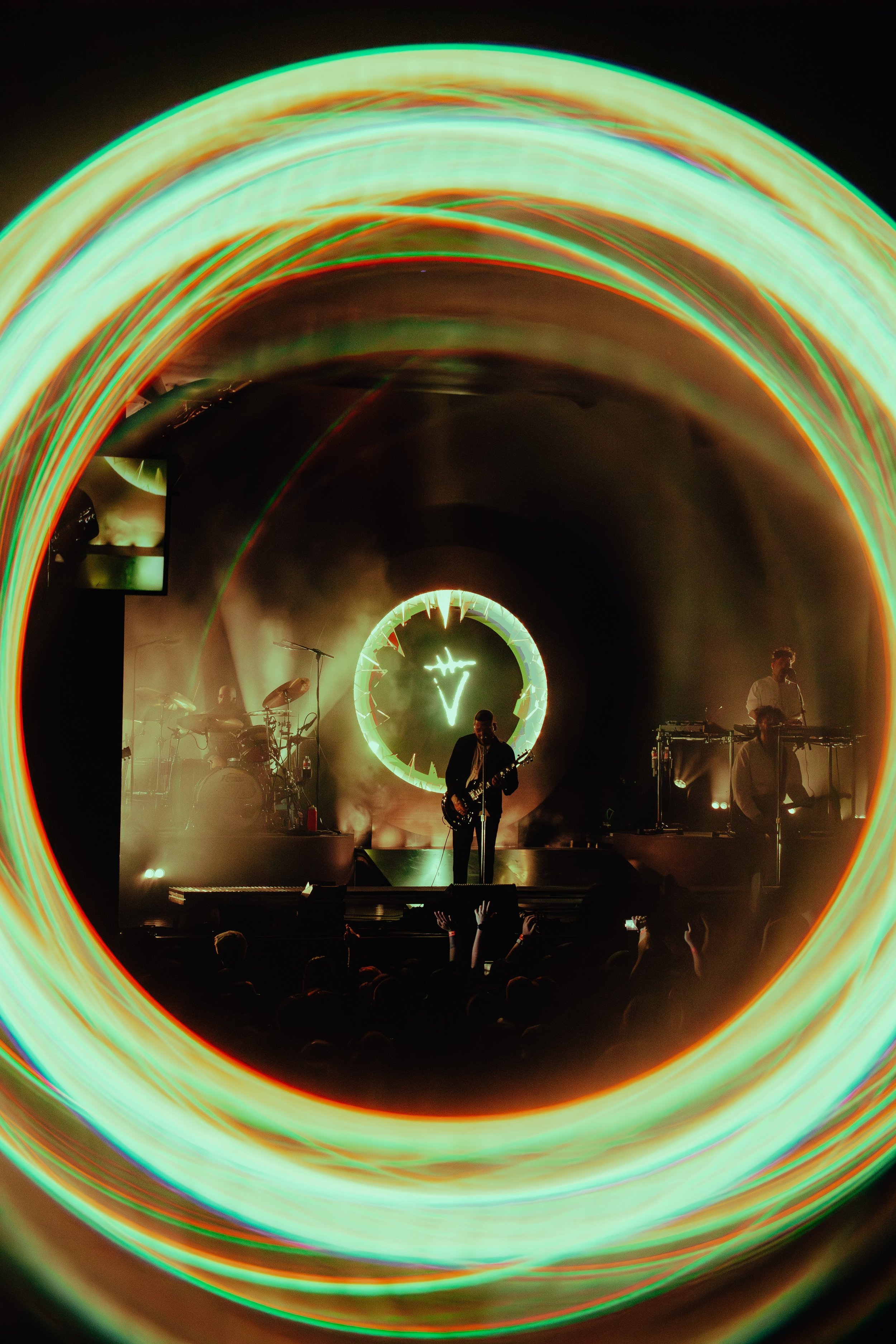 Live band performing on stage with a neon light logo in the background, surrounded by colorful light trails and an audience in the foreground.