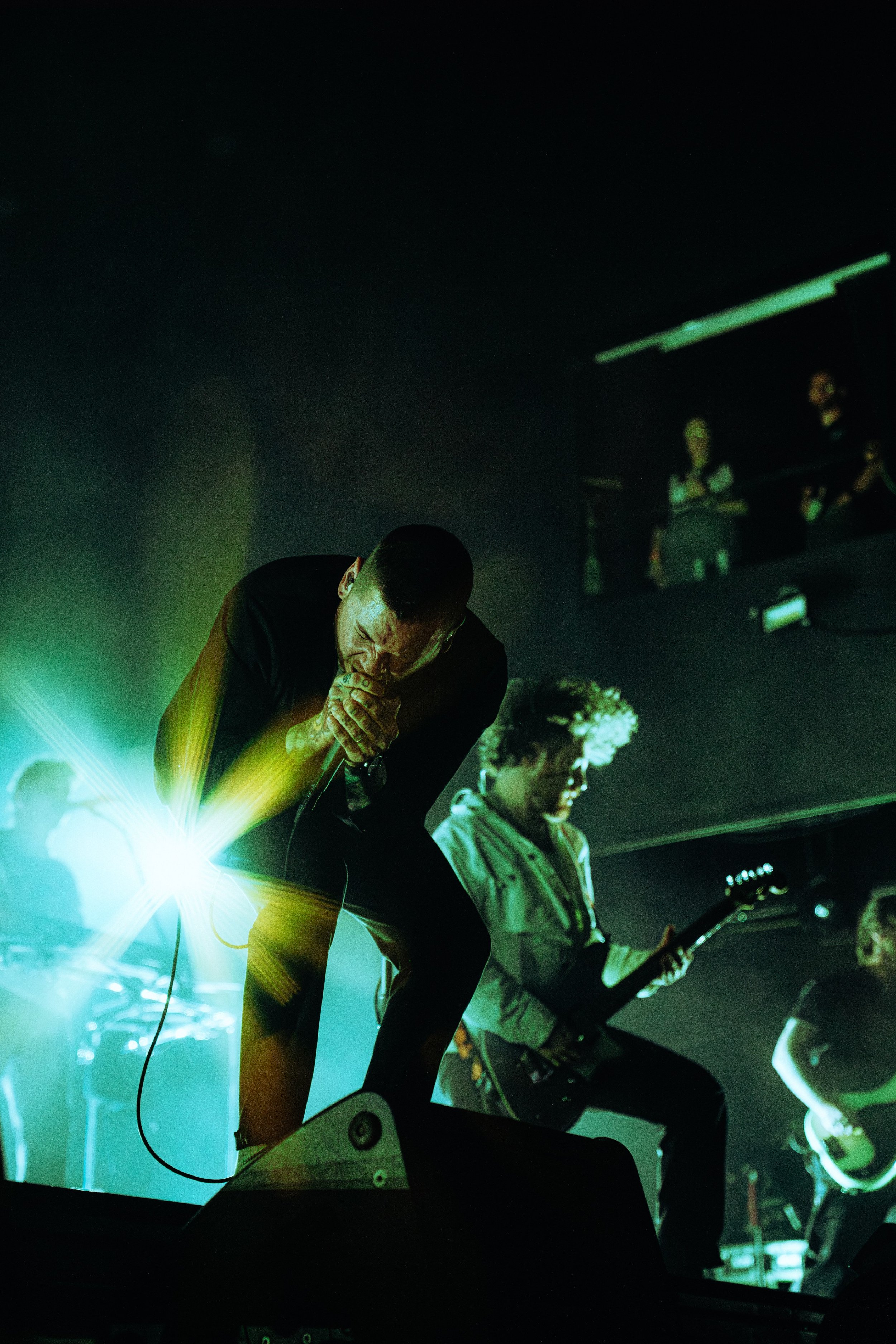 Musicians perform on stage with vibrant green and yellow lighting, one singer passionately singing into a microphone while another plays guitar.