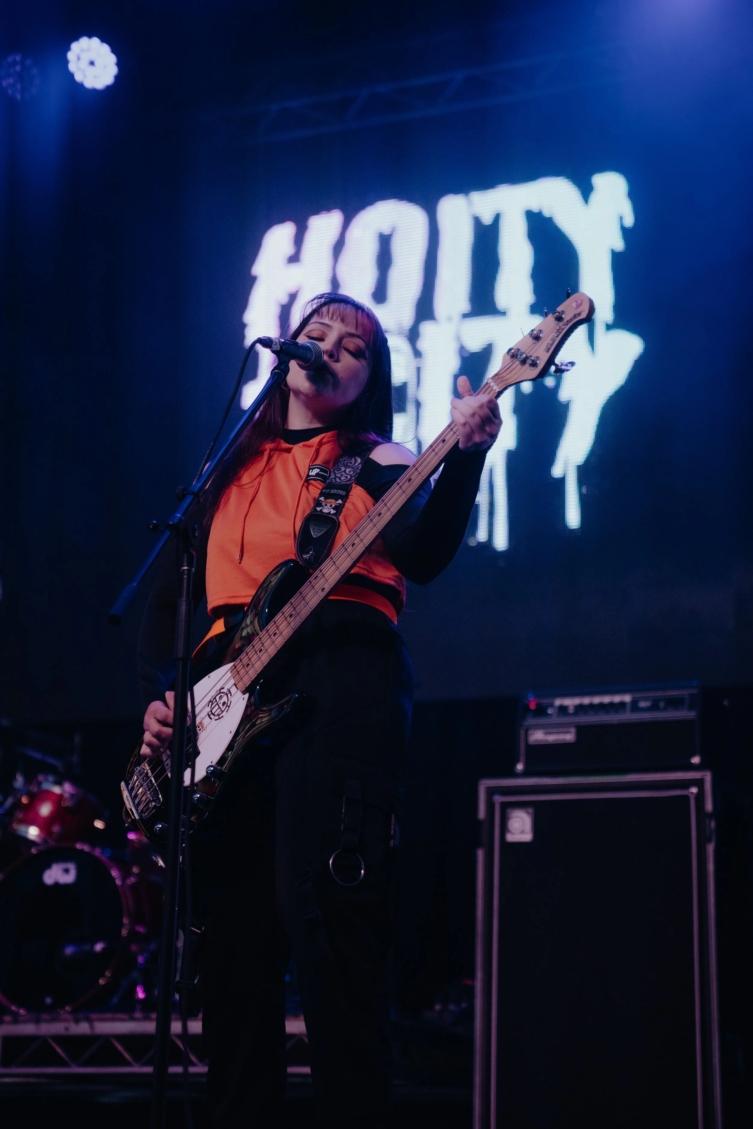 A woman singing and playing an electric guitar on stage with a blue background and a large illuminated sign that says 'HOUSE' behind her.
