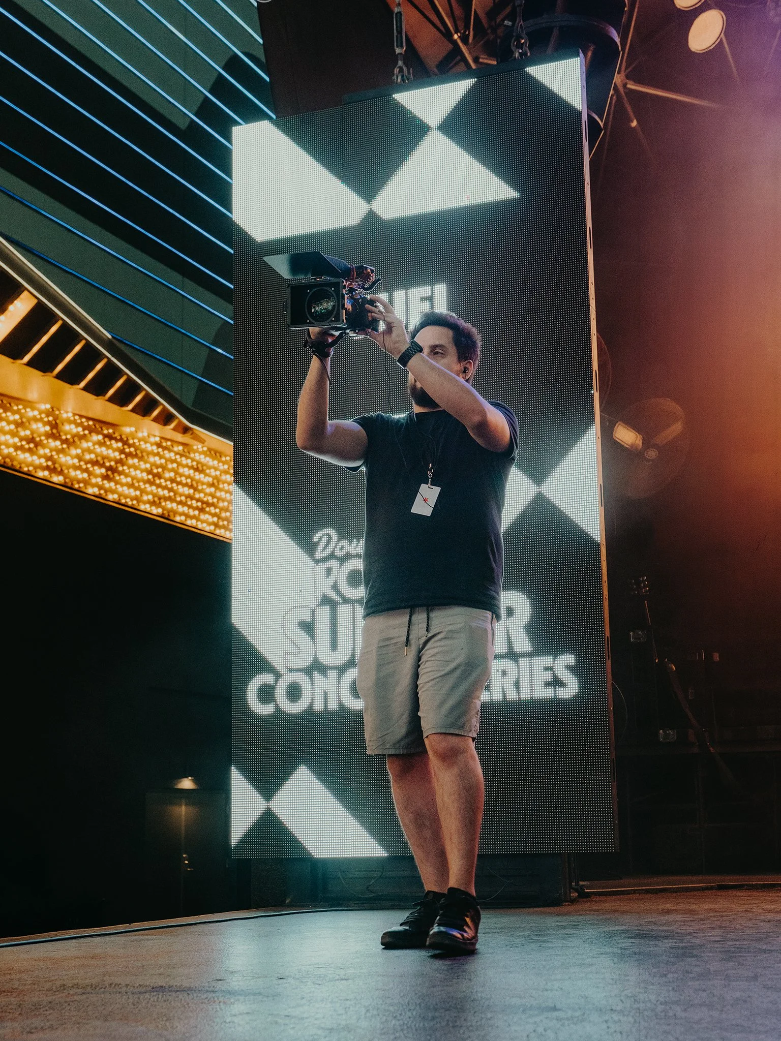 A man in a black t-shirt and shorts holding a video camera on a stage, with a digital screen in the background displaying the partially visible text 'DAVE CONNERS' and decorative lighting.
