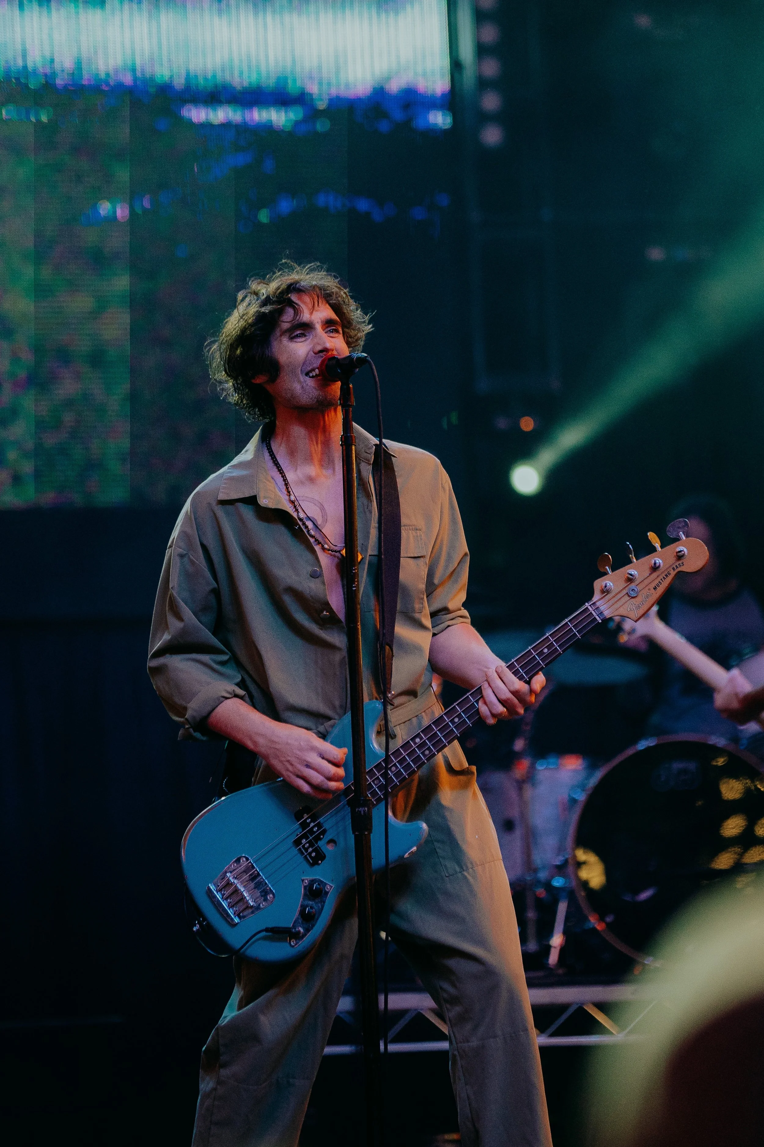 A person singing into a microphone while playing a light blue electric bass guitar on stage, with colorful stage lights and a large screen in the background.