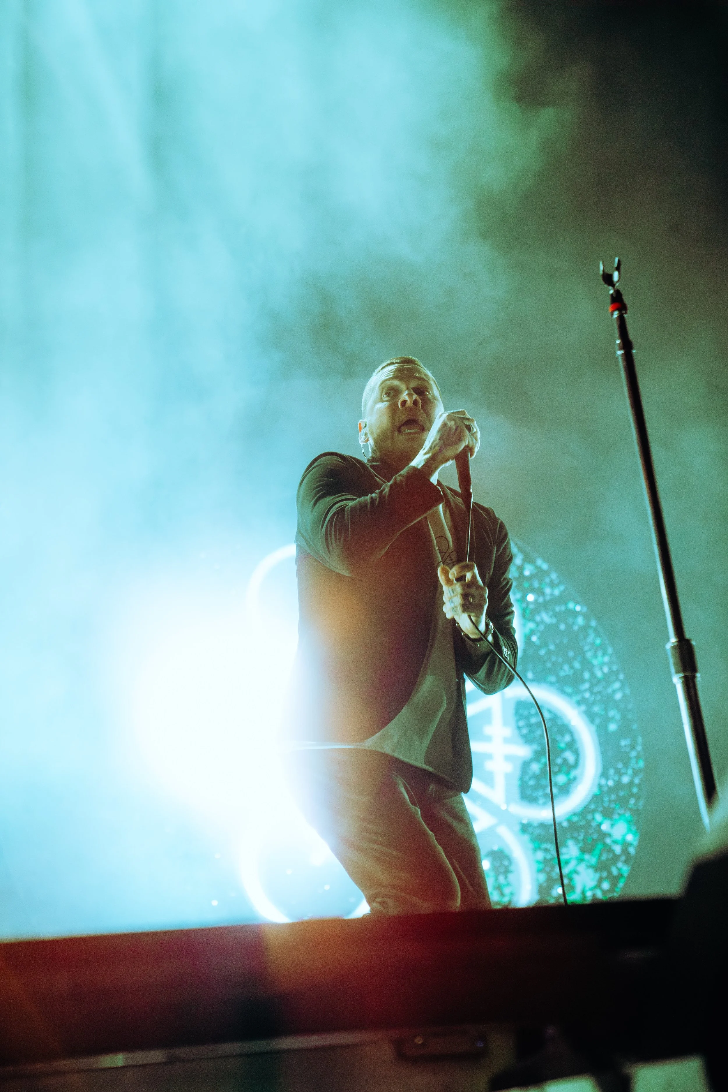 A man performing on stage, holding a microphone, with green and blue stage lighting and a large digital clock in the background.