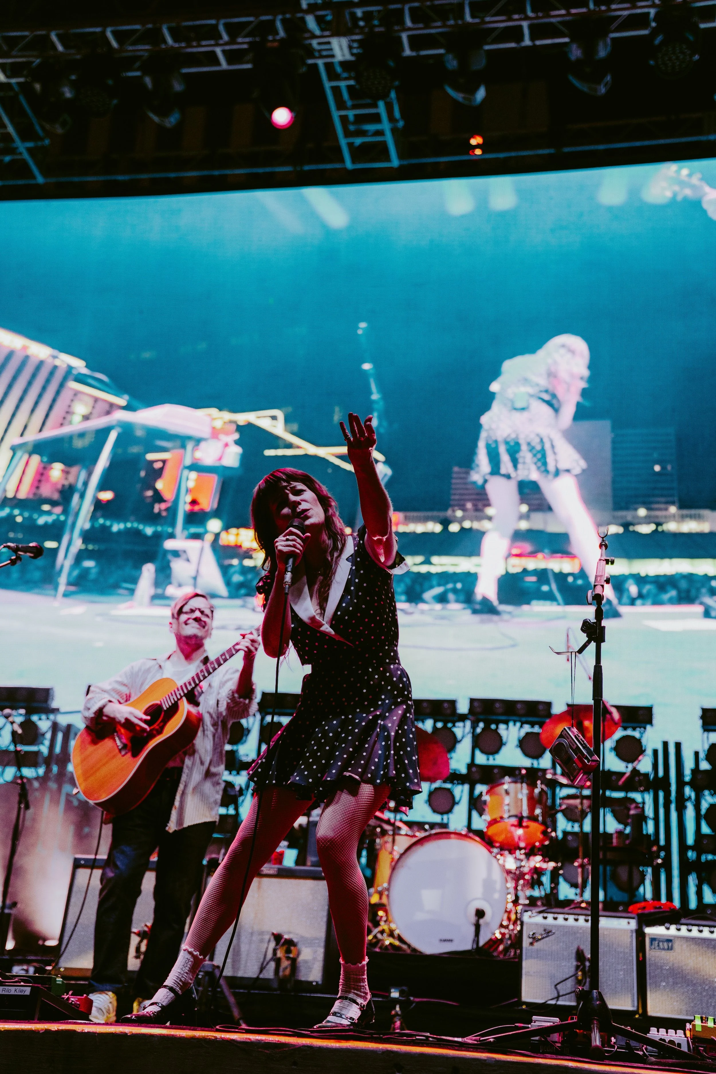 Performers on stage at a concert with a large LED screen in the background. One woman in polka dot dress and fishnet stockings singing into a microphone, a man playing guitar beside her, and a drummer in the background. Neon signs and city lights vis